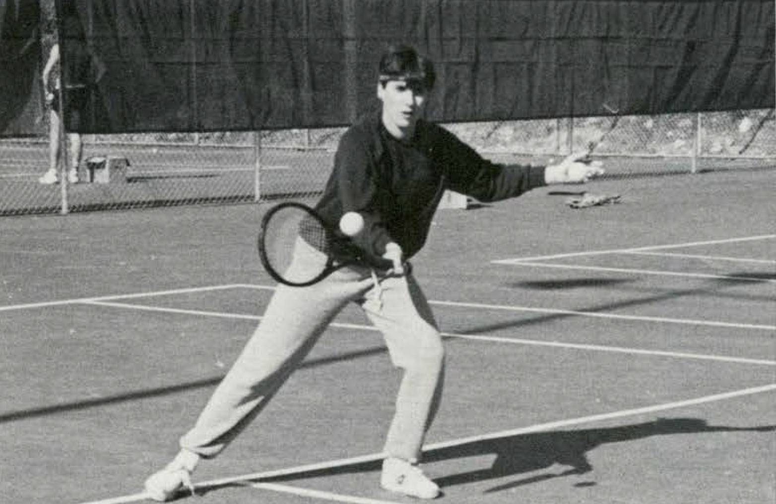 A black and white photo of a male student hitting a tennis ball with a racket while wearing a sweatsuit and a sweatband around his forehead. 