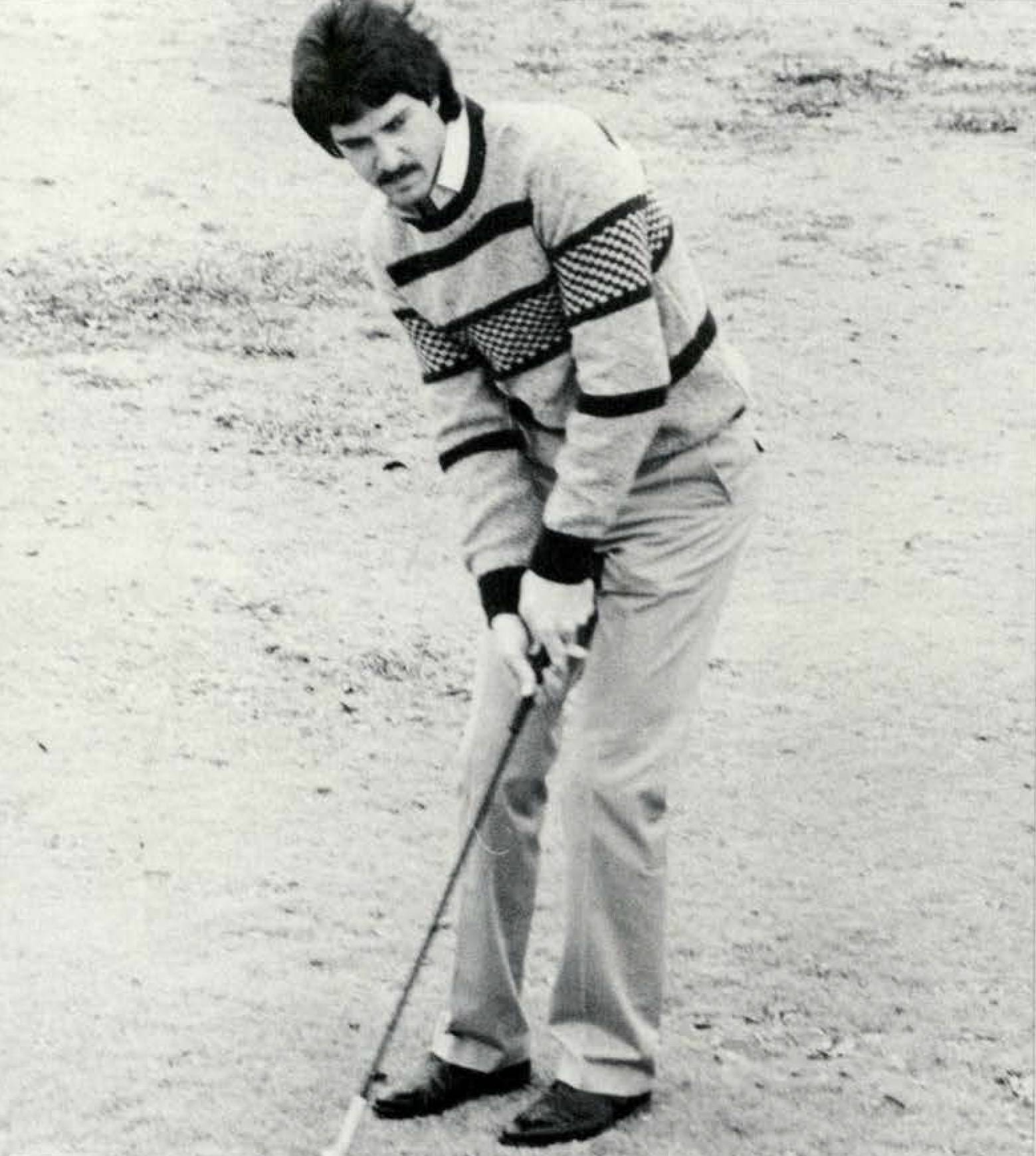 Black and white photo of a man golfing. He is wearing a button-down shirt, a striped sweater, and slacks. 