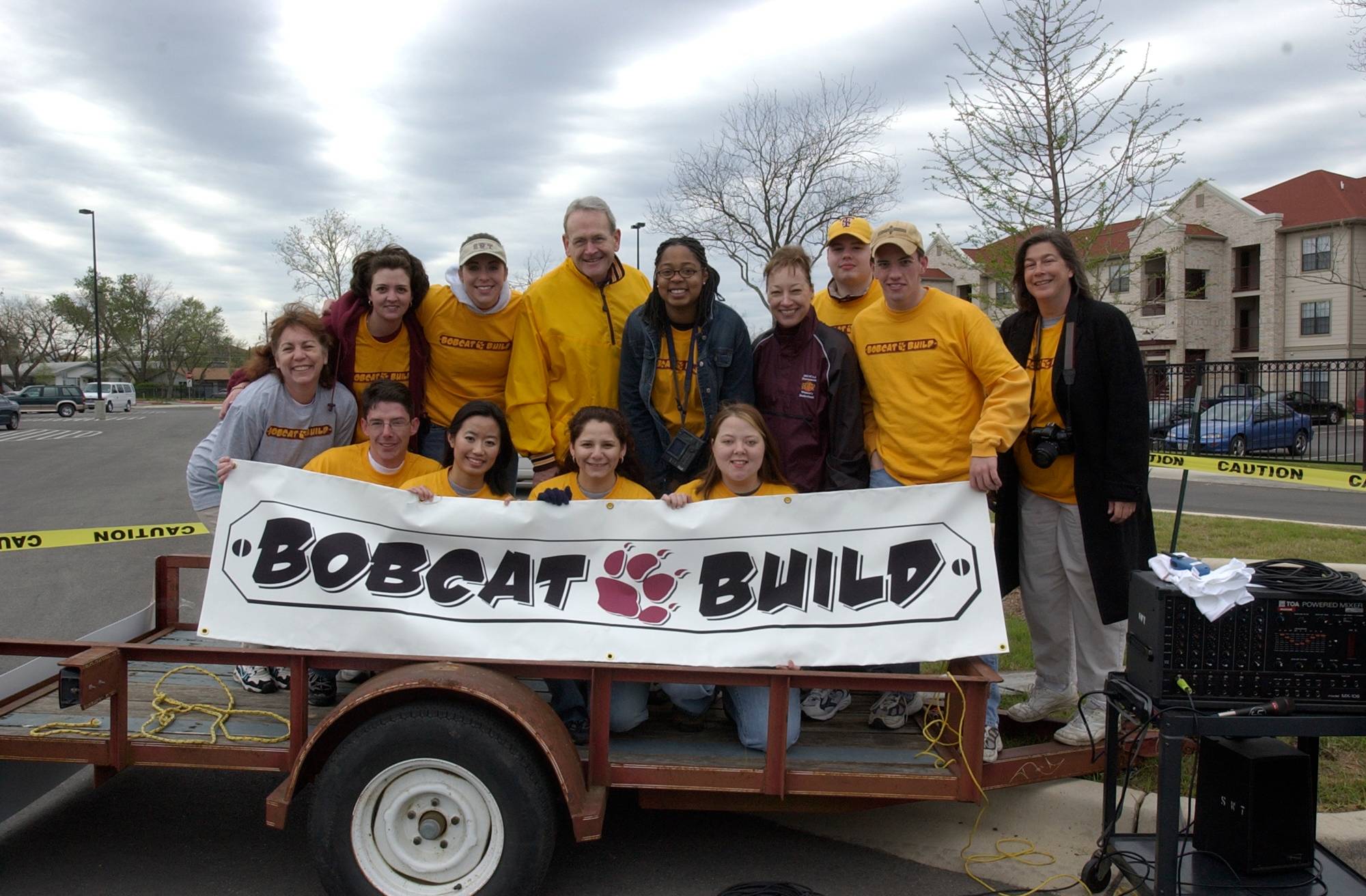 A group of TXST volunteers standing and smiling on top of a trailer, holding a banner that displays the Bobcat Build logo. 