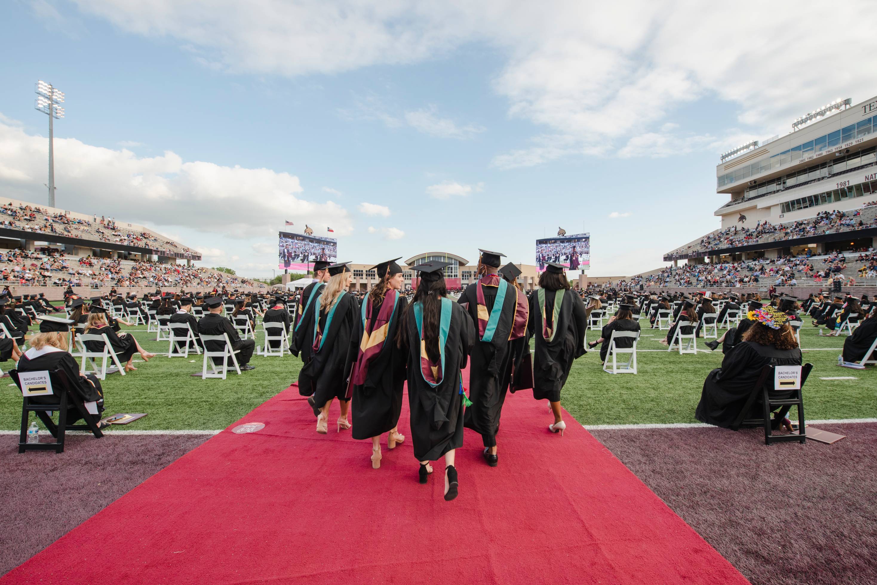 Seven graduates walking away from the camera during a commencement ceremony in a large outdoor stadium. People are sitting on the football field and in the stands. 