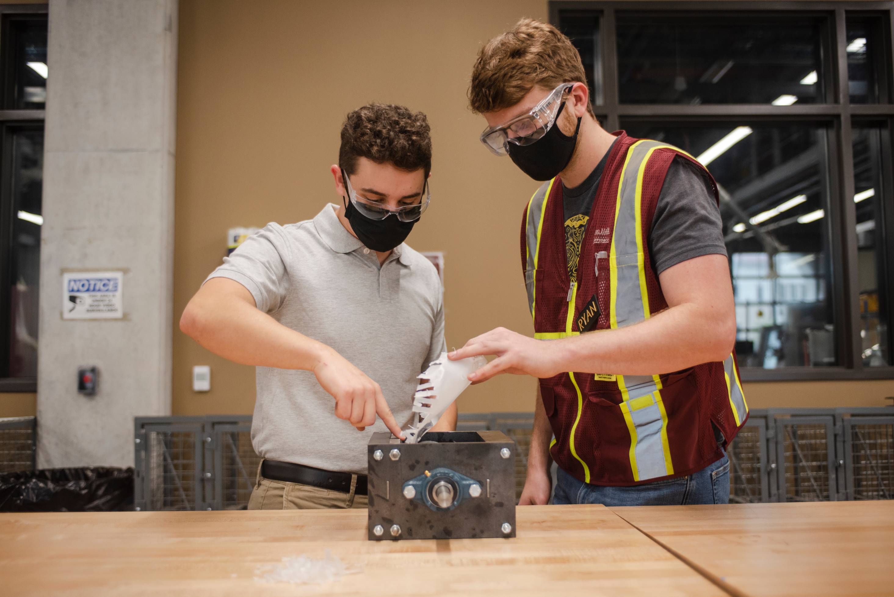 Two male students stand while working on a piece of machinery. They are wearing face masks and safety glasses. 