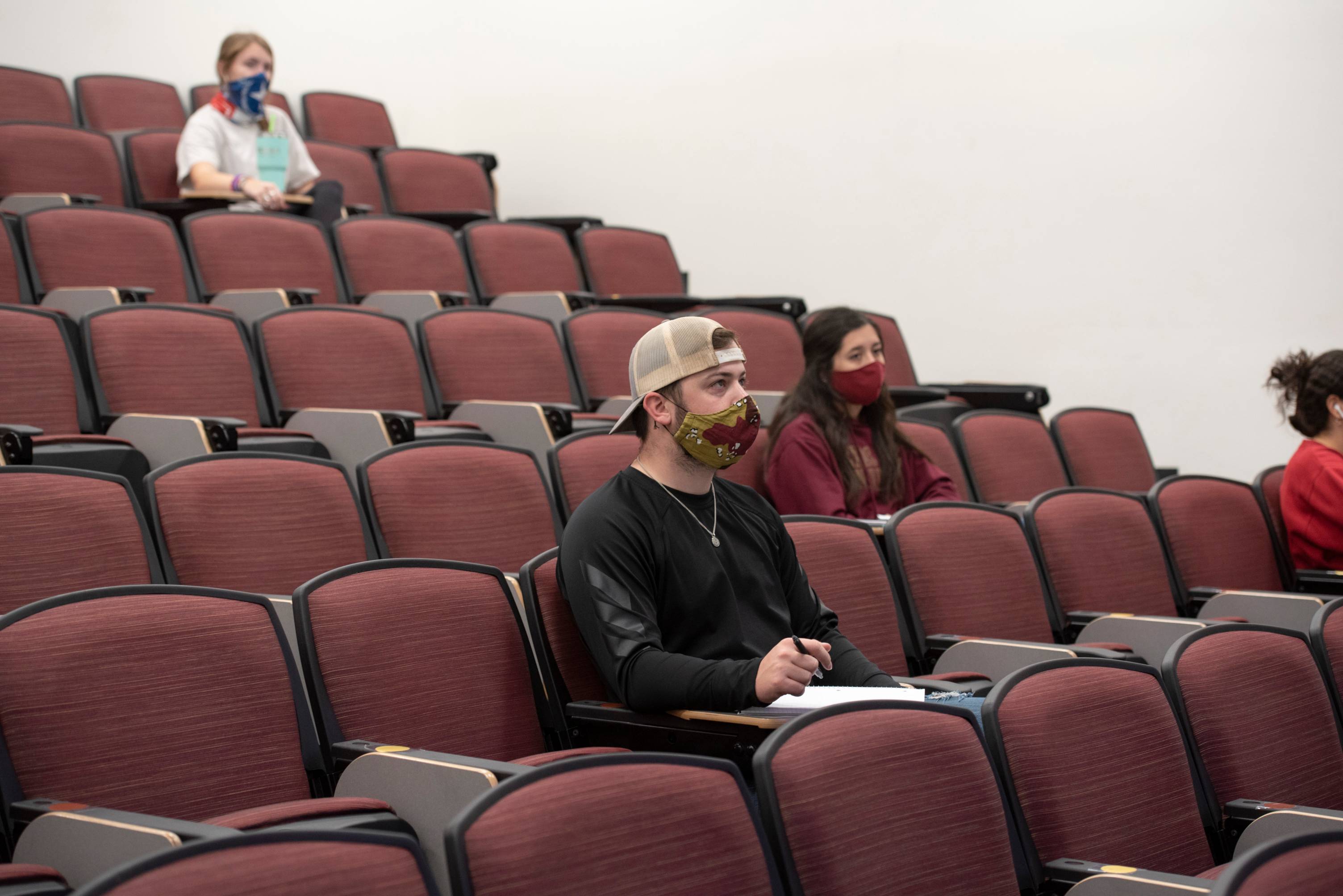 Four students are sitting in a stadium-style classroom with maroon seats. The students are wearing face masks and practicing social distancing. 