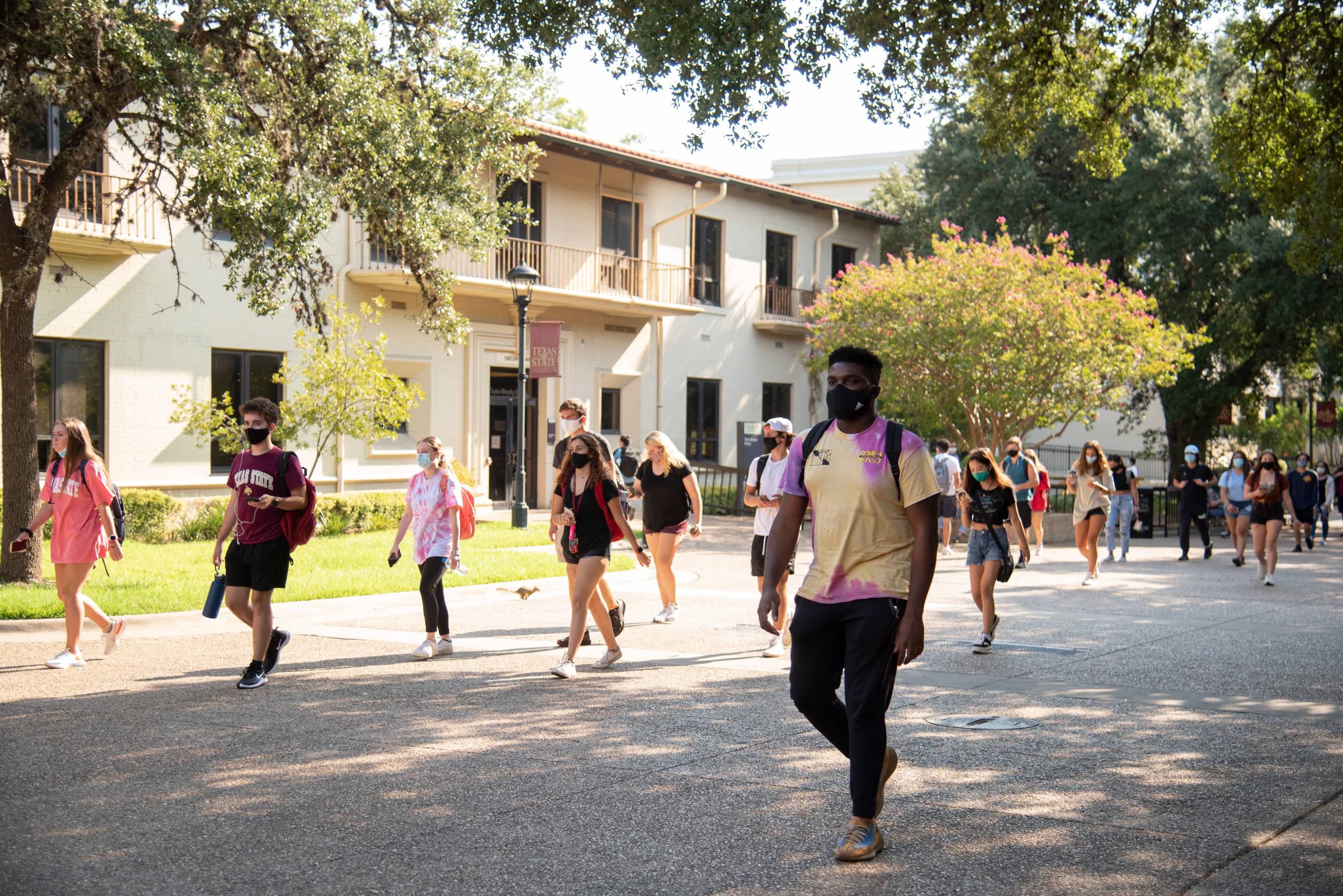 A walkway between campus buildings is filled with students walking in one direction with face masks on. 