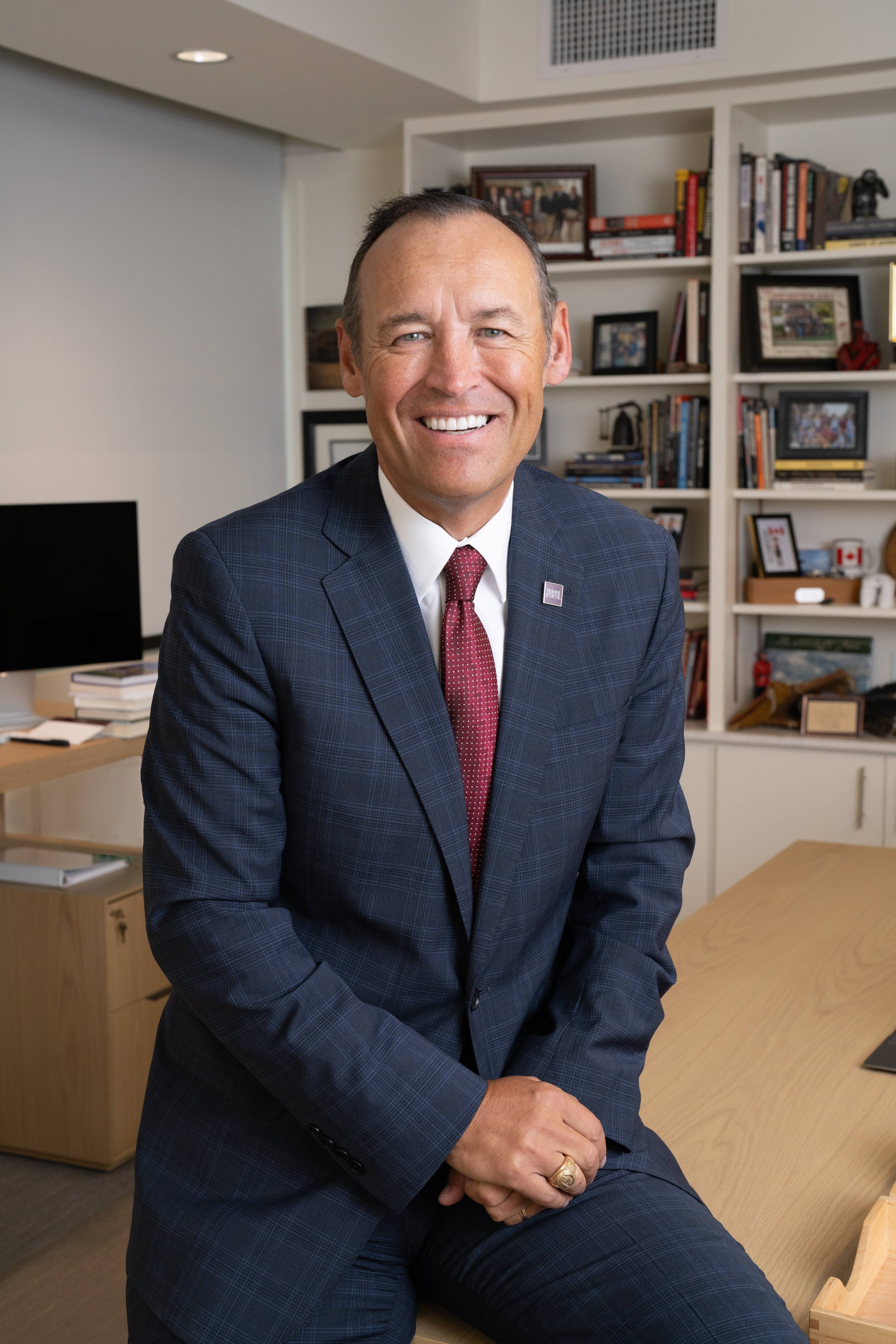 TXST President Kelly Damphousse is seated in his office, wearing a striped blue suit, white shirt, and maroon tie. His hands are placed on his lap. The background is adorned with personal photos and a selection of books.