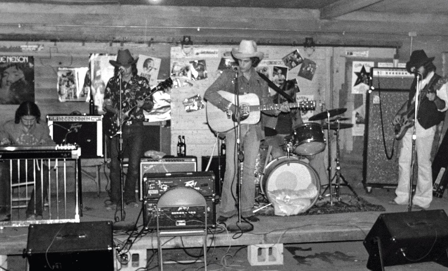 George Strait and other band members appear to be preparing for a performance as they gently strum their guitars. A pianist sits to the right of the stage and a drummer in the background as George is flanked by two fellow guitarists. Amplifiers and speakers can also be seen spread about the stage. 