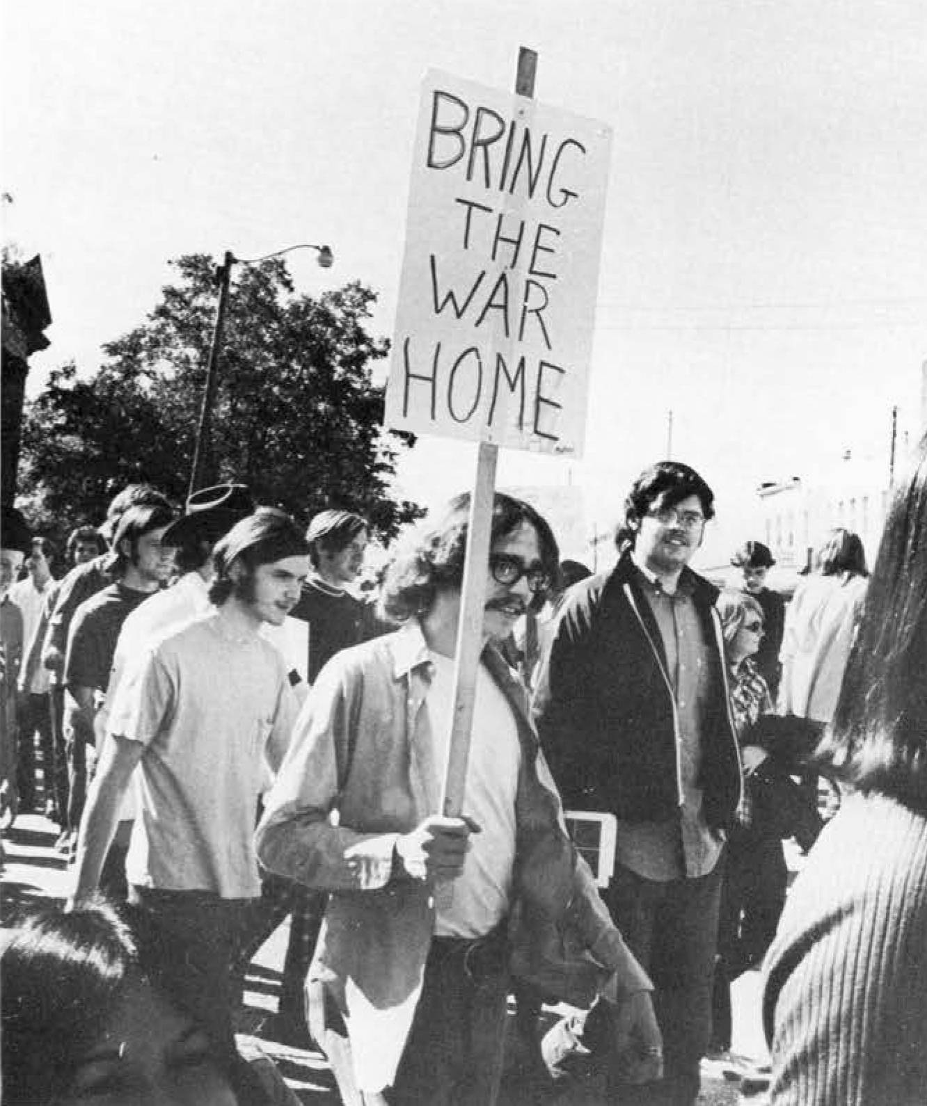 Black and white photo of a bespectacled young man in the 1960s holding a sign that reads "Bring the War Home". Behind him are other young men and women engaged in a protest march.