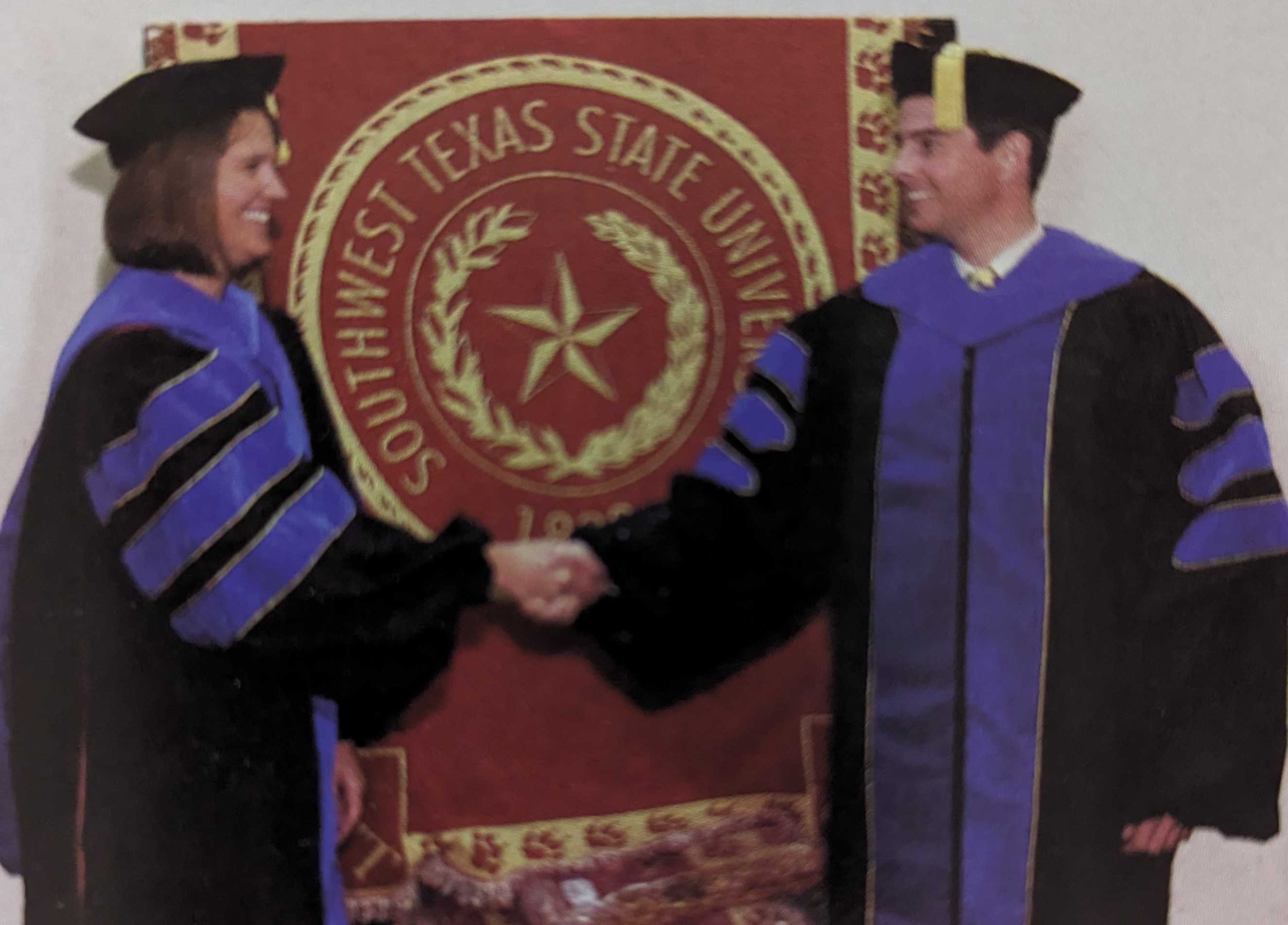 Dr. Lisa DeChano and Dr. Todd Votteler shake hands in front of a Southwest Texas State University banner at their Ph.D. graduation ceremony. Each wears a black robe trimmed in blue down the front, with blue bars down the sleeves. 