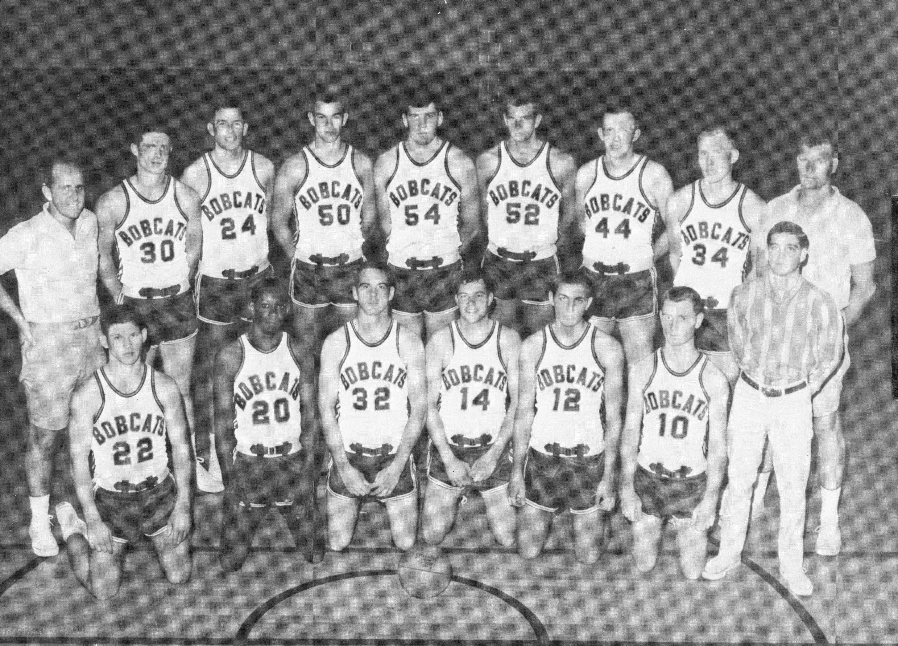 Black and white photo of young men in 1960s basketball uniforms posing in two rows, flanked on either side by coaches. The back row stands and the front row kneels. Johnny E. Brown is kneeling and positioned second from the left, the only Black person in the image.