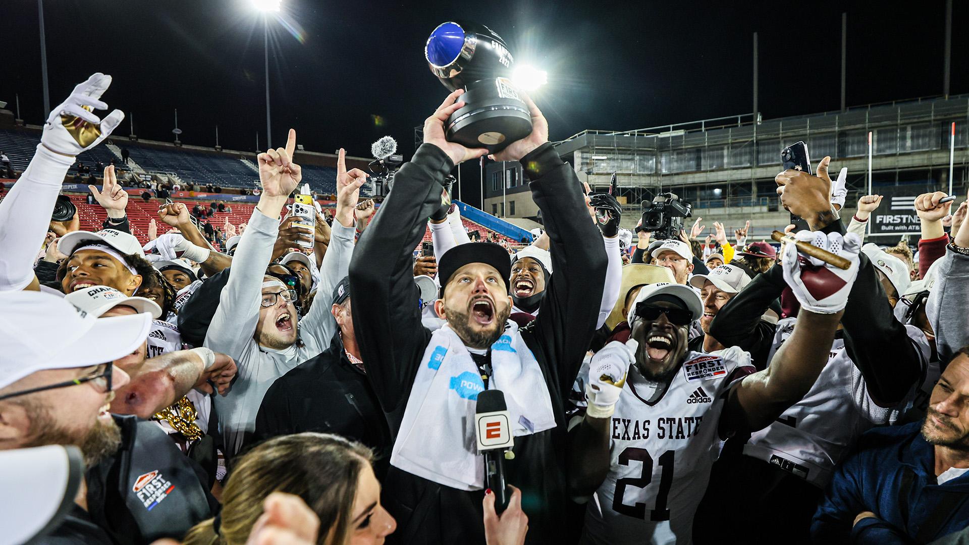 Coach GJ Kinne lifting the SERVPRO First Responder Bowl Trophy with the TXST Bobcat Football team cheering in celebration around him. 