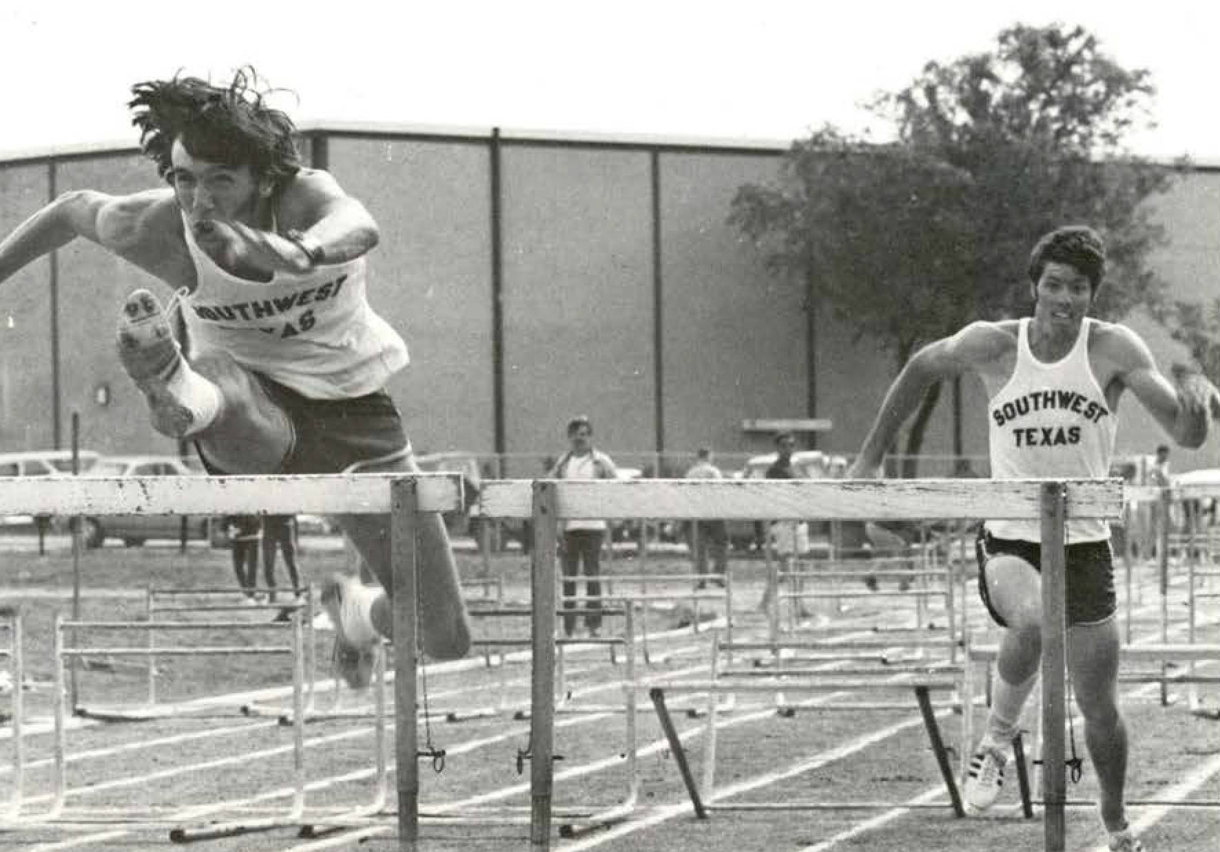 Two Southwest TXST track athletes competing in a hurdling race. One athlete is mid-hurdle, the other is approaching his next hurdle. 