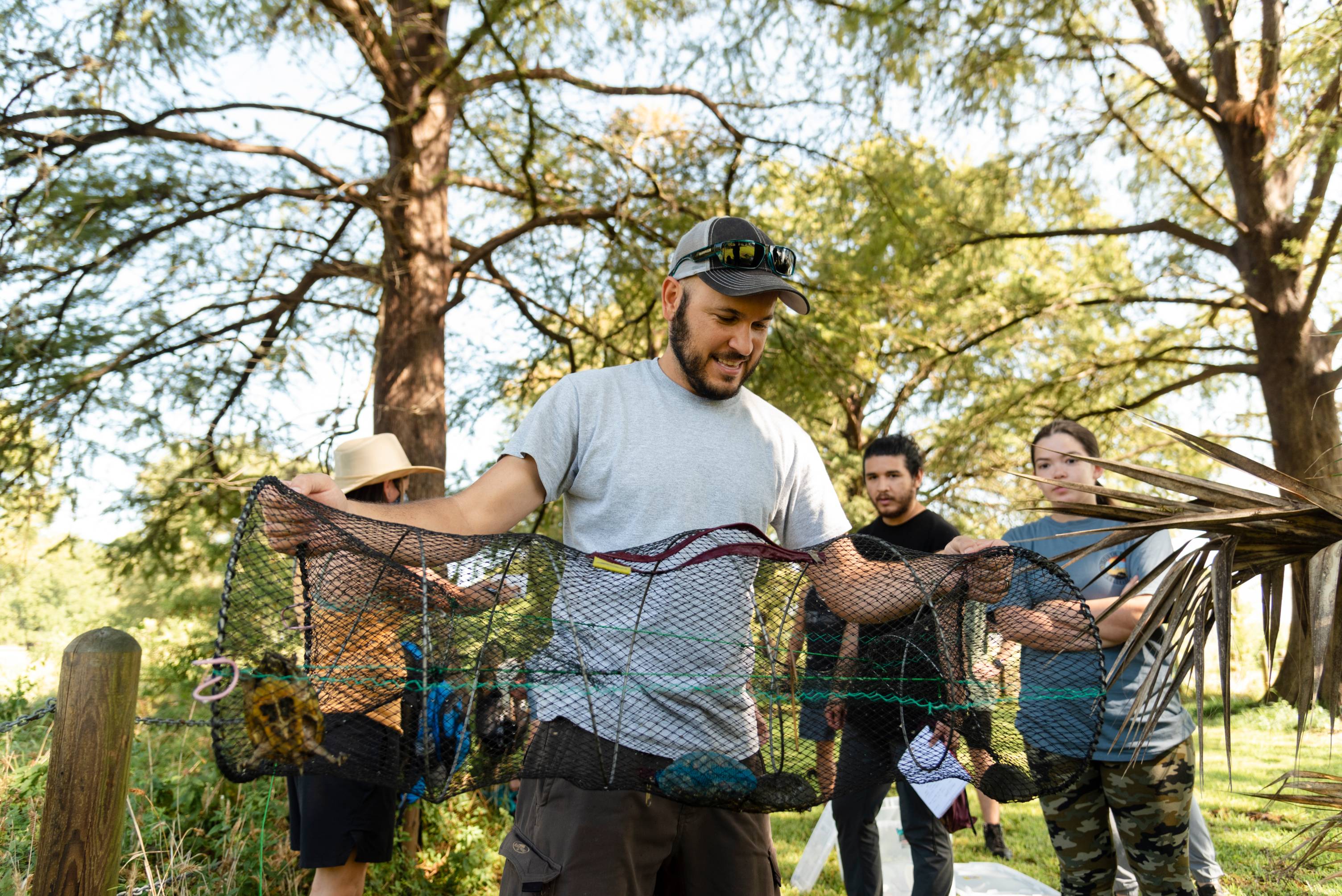 Students and researchers outside the Meadows Center engaged in a study on the local turtle population. A young man holds up a turtle trap containing several specimens, while others look on in interest.