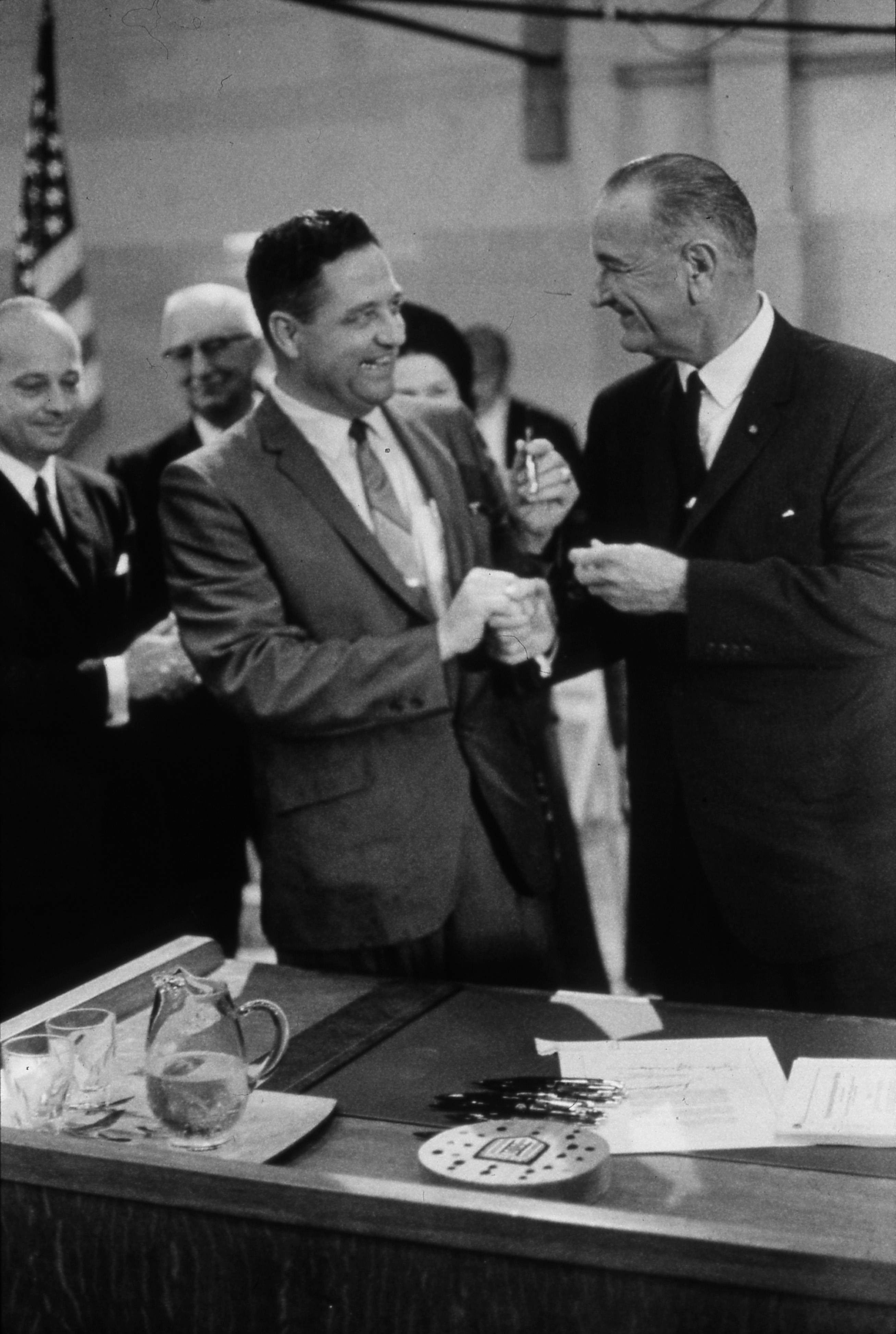 Black and white historic shot of Lyndon B. Johnson, dressed in a suit, shaking hands with another man, who is holding a pen. Both men are smiling and before them is a desk littered with documents. Several people in suits and an American flag stand behind them.