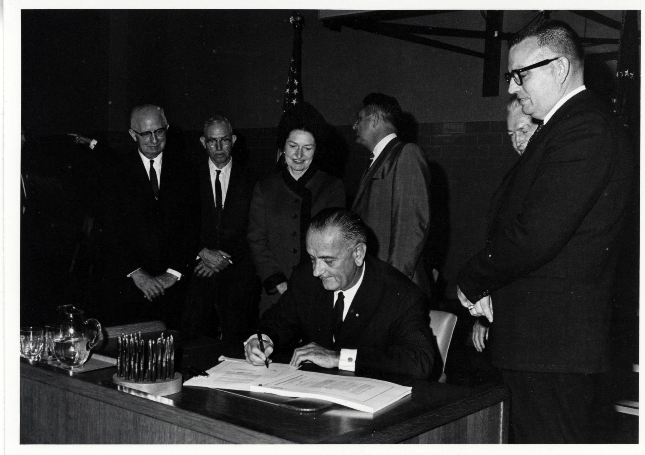 Dimly lit black and white photo of Lyndon B. Johnson sitting at his desk, signing a document. Behind him are several men and one woman in suits. Their hands are clasped as they're intently watching the president.