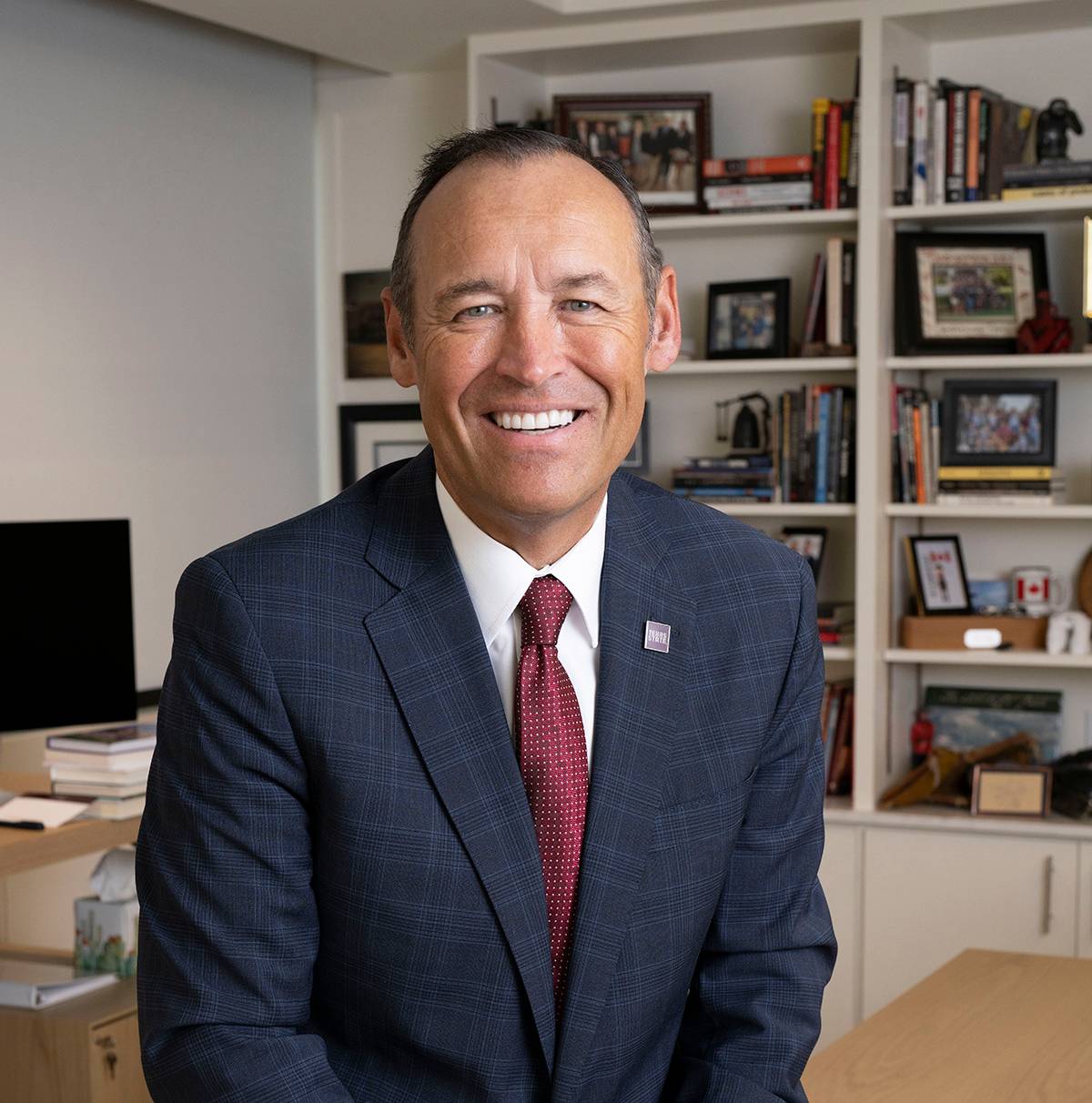 A smiling man in a suit posing for the camera. Behind him is a bookshelf full of books.