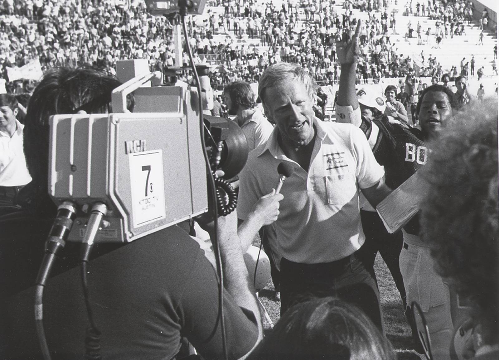 Black and white photo of a football coach being interviewed by a news station. Behind him is a football player holding up a finger in a gesture that denotes their "number 1" status. Behind them is a crowded football field and spectator stands full of people.