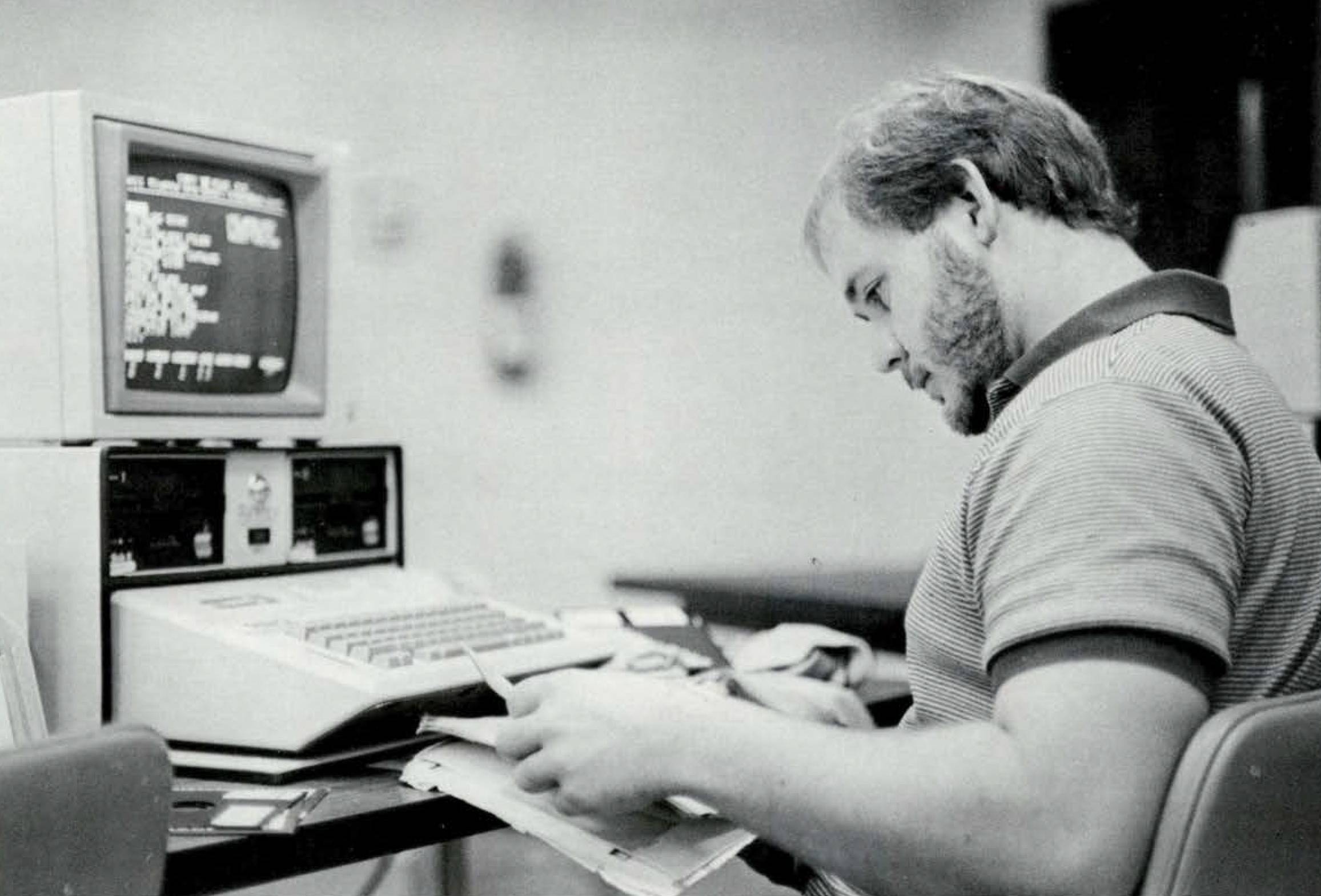 Black and white photo of a young man in a polo shirt, flipping through a book. In front of him sits a retro computer, displaying lines of code.