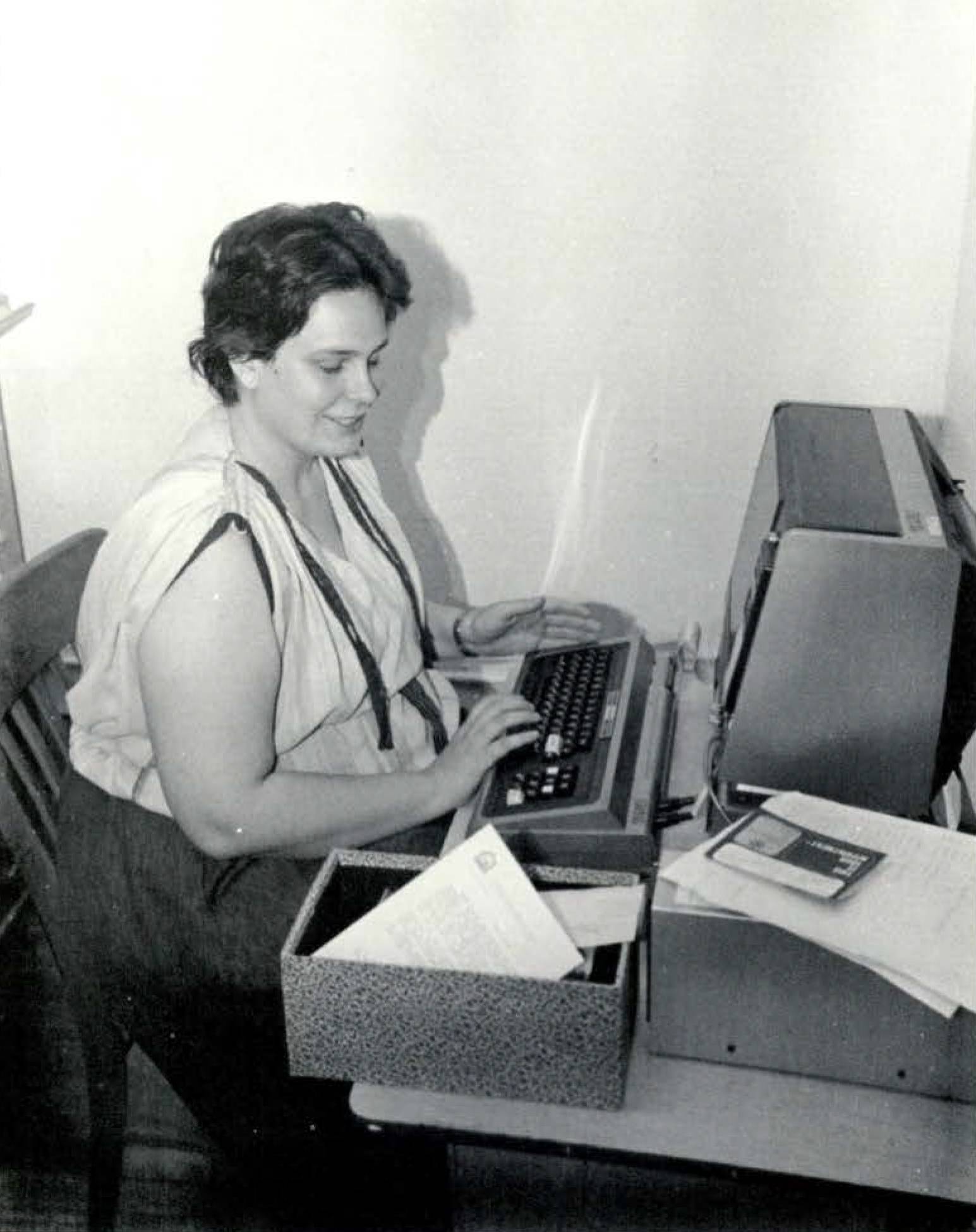 Black and white photo of a young woman smiling faintly as she types on an early computer.