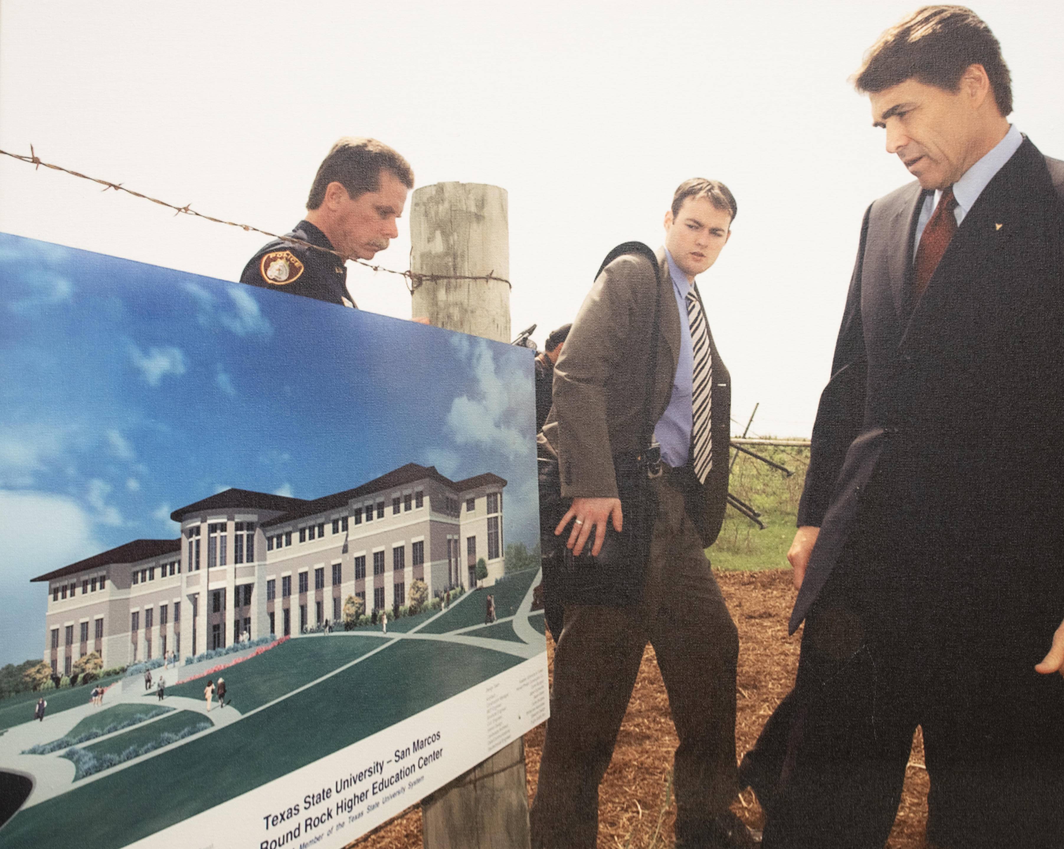 Former Governor Rick Perry, wearing a dark suit and accompanied by a uniformed officer and another man in a suit, looks at a poster displaying a building announcement. The poster has an image of a building and text that reads: "Texas State University - San Marcos -- Round Rock Higher Education Center"