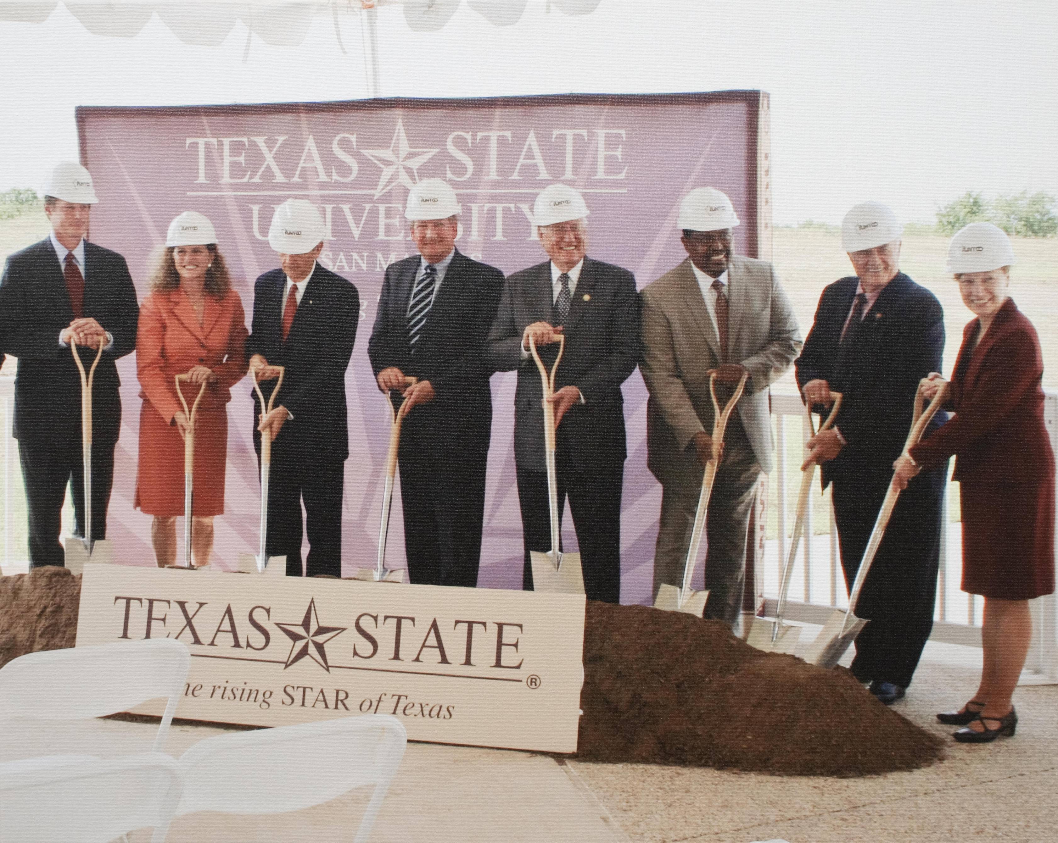 A group of men and women in suits and pencil skirts wear hard hats and pose proudly in front of a pile of dirt. They each hold a shovel. In front of them is a sign that reads: "Texas State -- The Rising STAR of Texas"