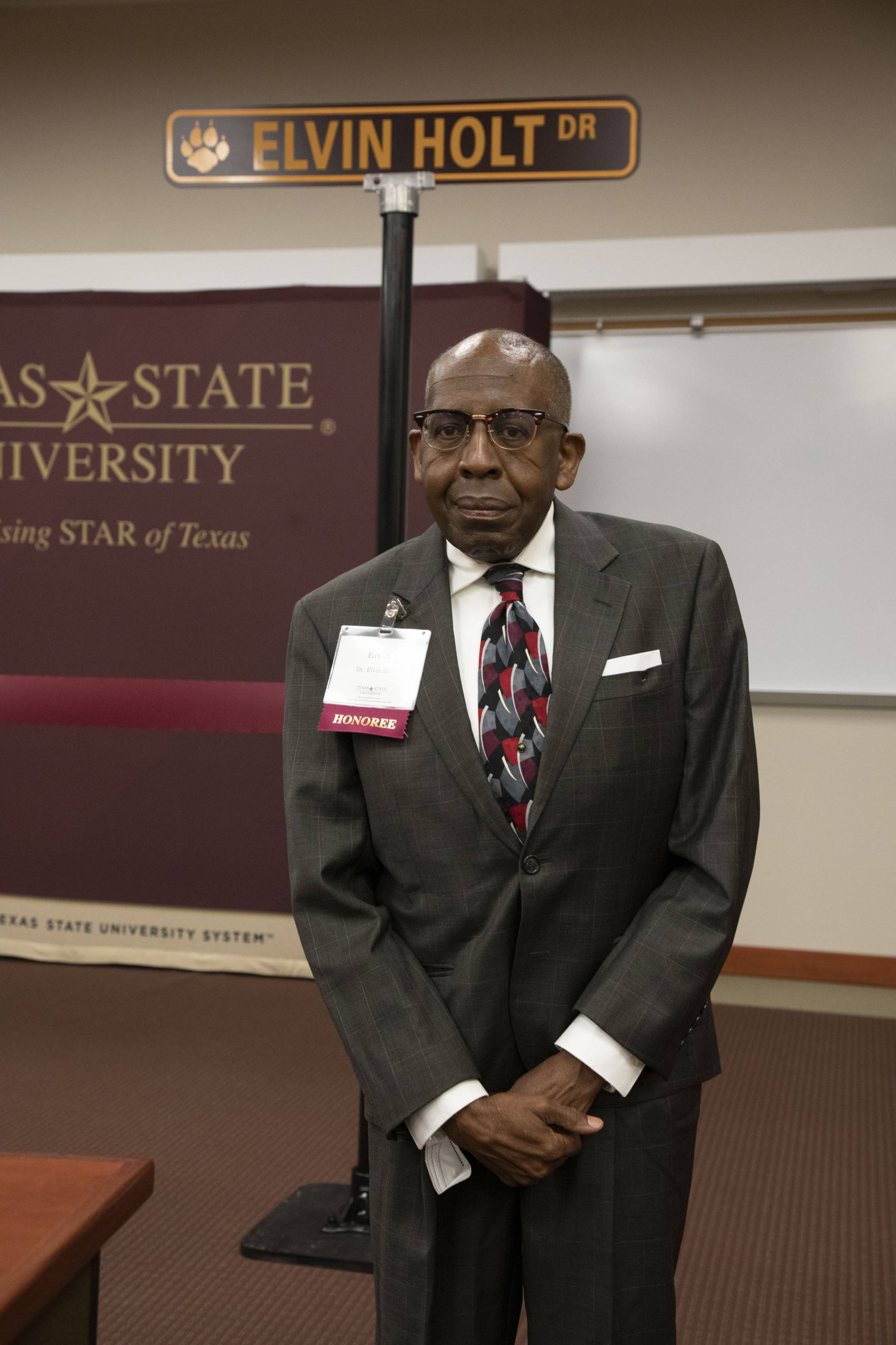 Photo of a man in a suit wearing retro glasses and clasping his hands. Behind him is a street sign that reads: "Elvin Holt Dr"