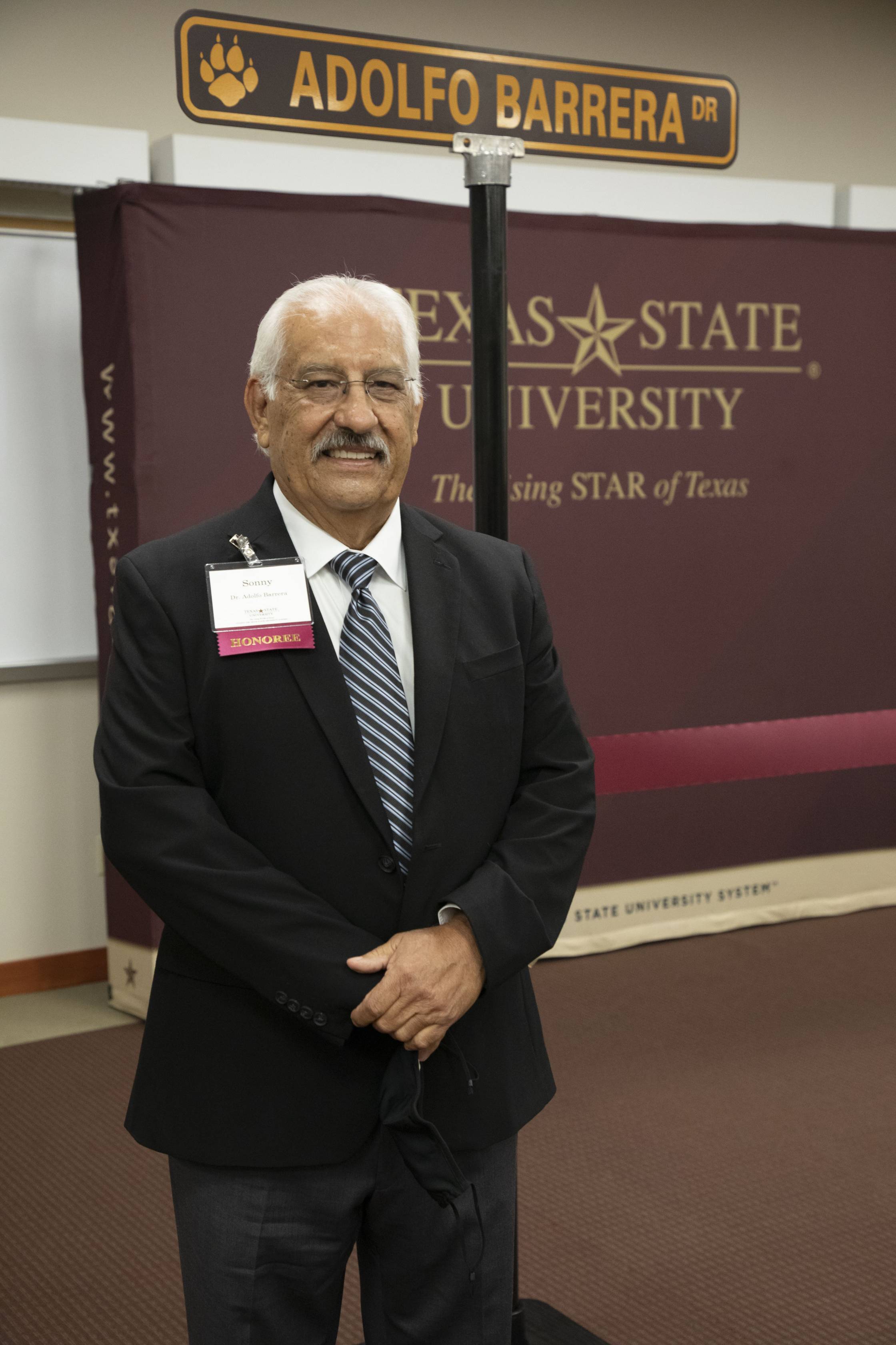 Photo of a man in a suit smiling in front of a sign that reads: "Adolfo Barrera Dr"