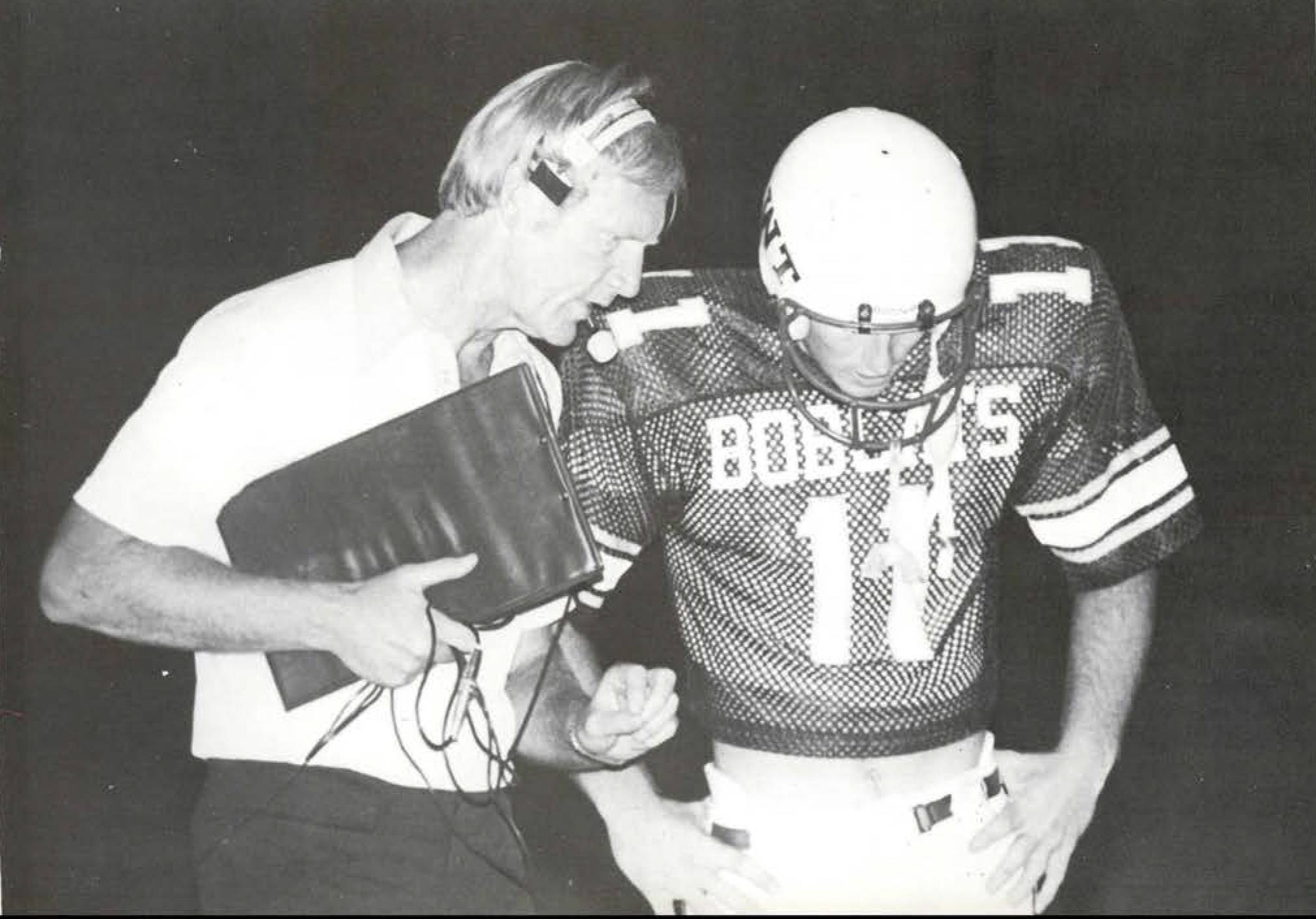 Coach Jim Wacker is seen in profile, in the midst of explaining a play to a football player. The coach wears headphones and carries a clipboard with one hand, gesturing with the other hand. The football player wears a jersey emblazoned with "Bobcats 11" and stands with his hands on his hips.