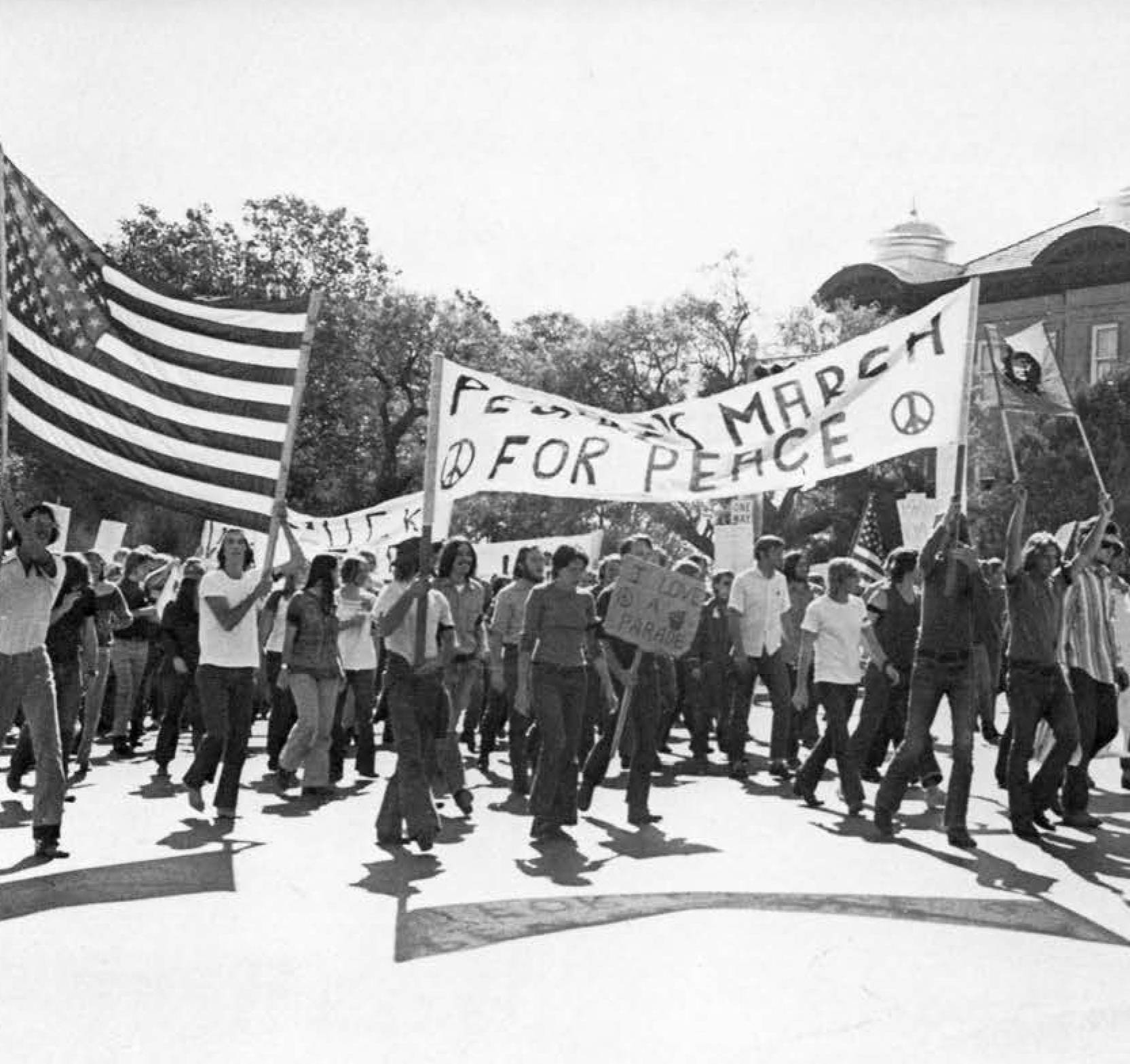 Black and white 1970s photo of crowd marching in the street. Prominently held aloft is a homemade banner, reading "March for Peace" with two peace symbols drawn on. Behind them are smaller banners and an American flag hoisted high by protesters. 