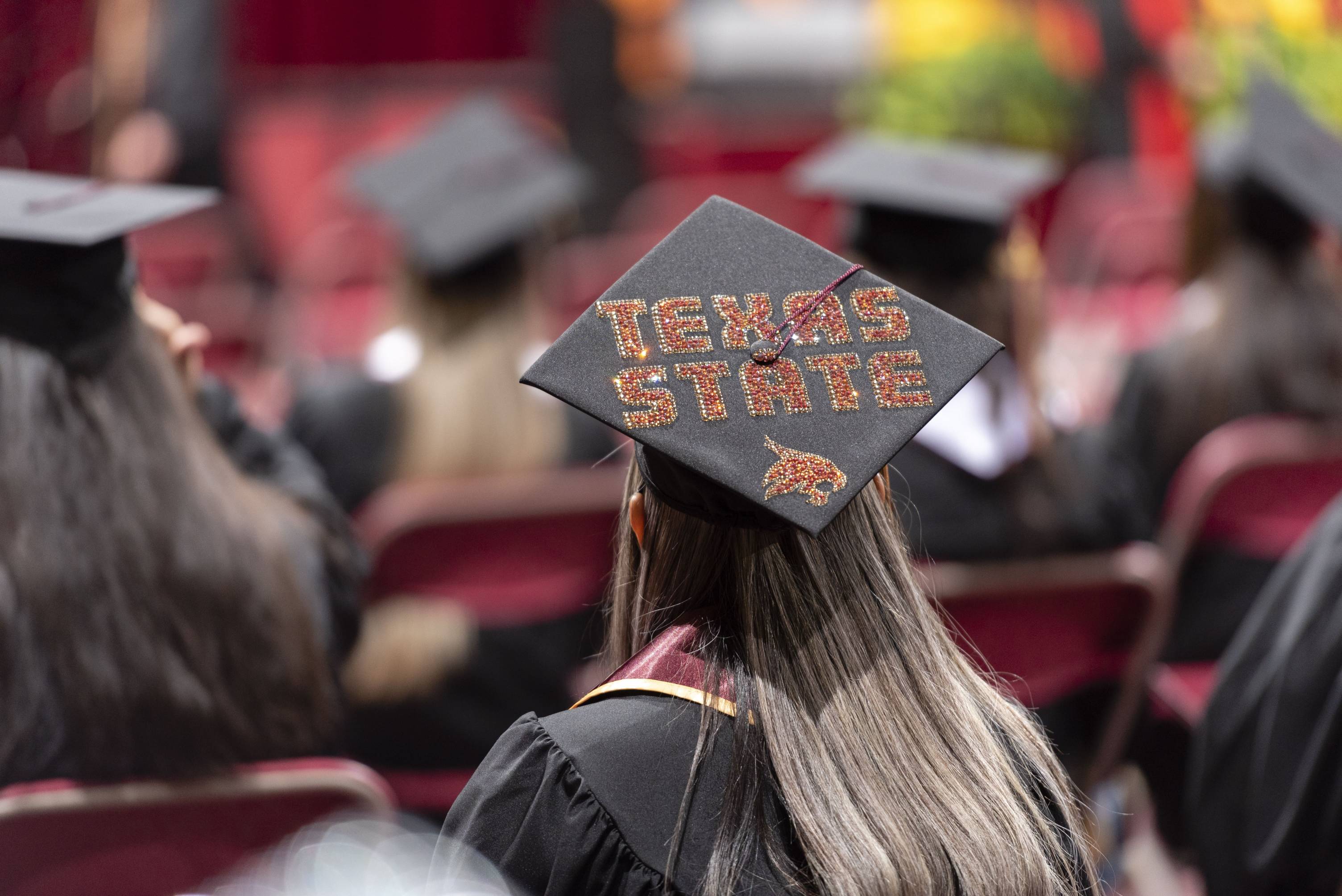 A photo of the back of a seated girl with long hair. She wears a graduation gown and cap. The cap is bedazzled with "Texas State" on top of it.