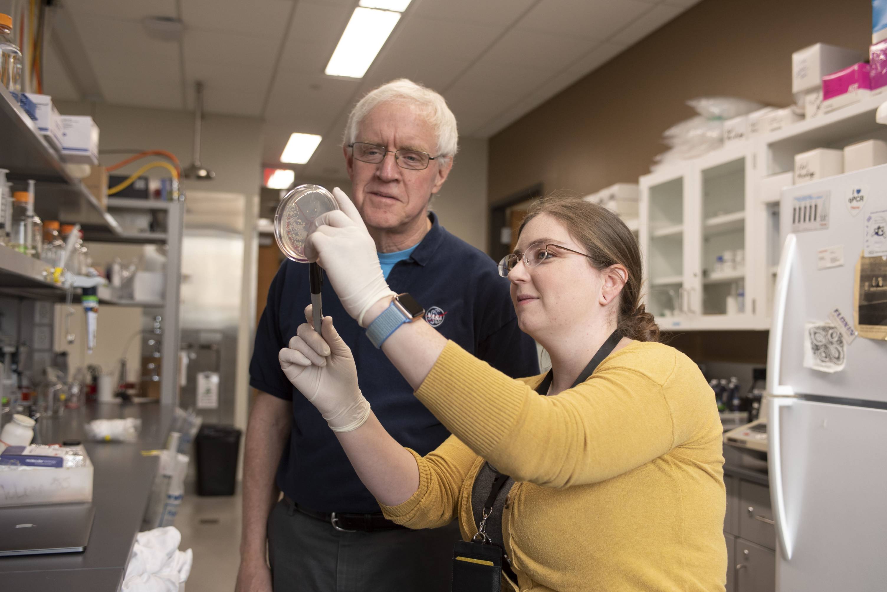 A young woman in a yellow cardigan holds up a petrie dish for an older man to inspect. Lab instruments can be seen in the background.