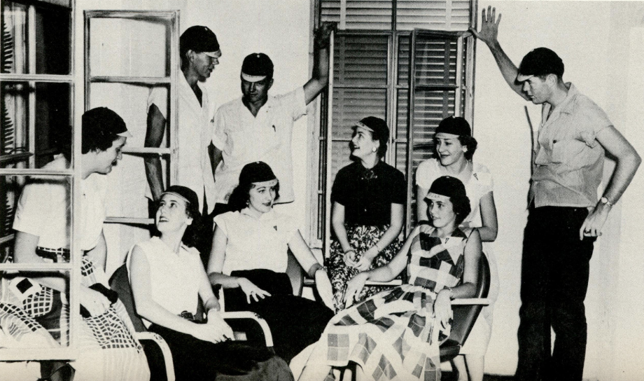 Black and white snapshot of a group of nine freshmen co-ed students mingling on the porch of a building. Each wears the traditional freshman beanie.