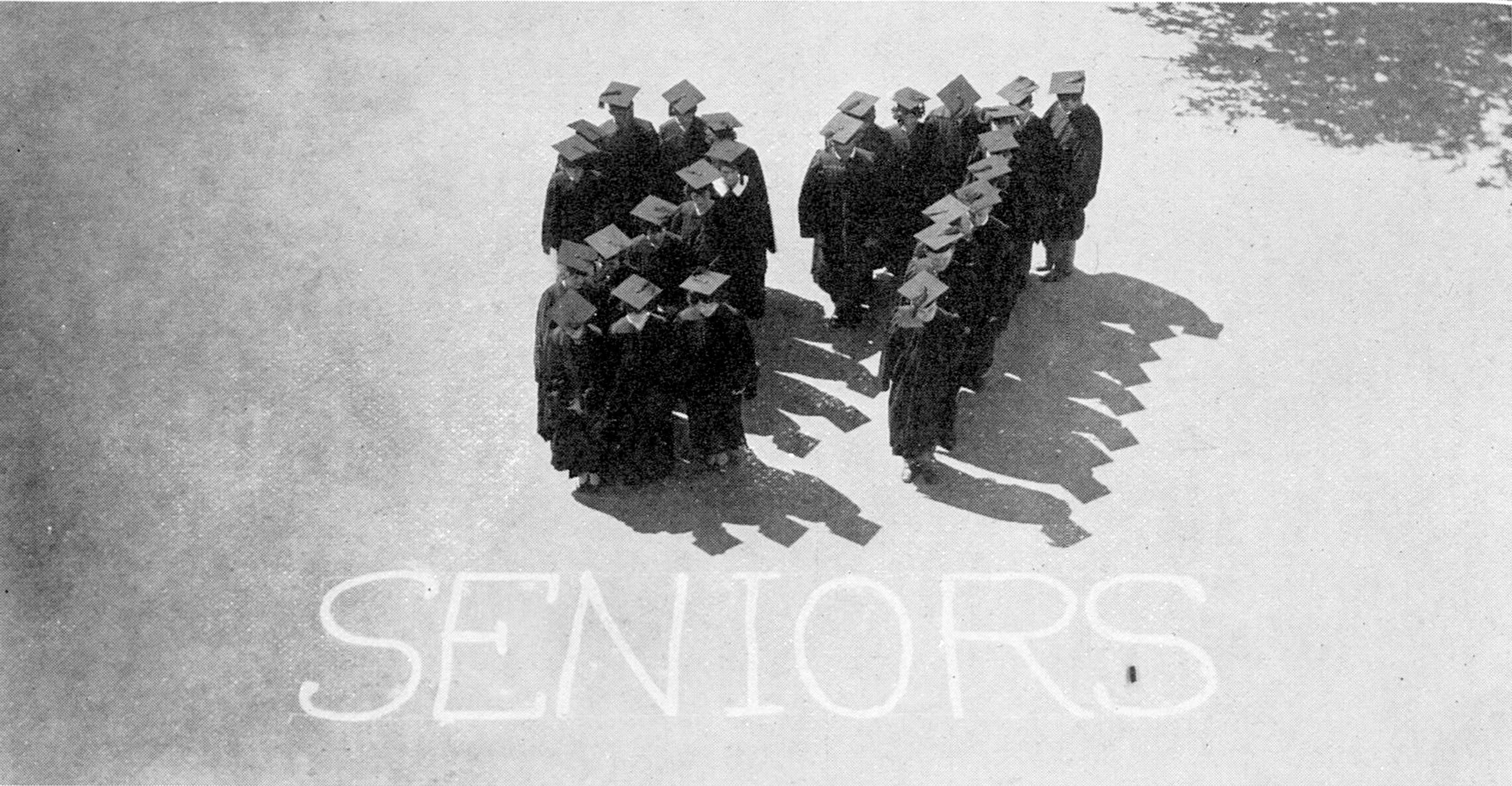 Aged black and white photograph of graduating seniors in 1927. The photo is taken from above, and the students stand in lines in dark robes and mortarboards with their backs to the camera. They've lined themselves up so that when viewed from above, their queues spell '27.' Beneath their two lines, they've written the word 'seniors' in chalk, so the photograph reads '27 Seniors.'