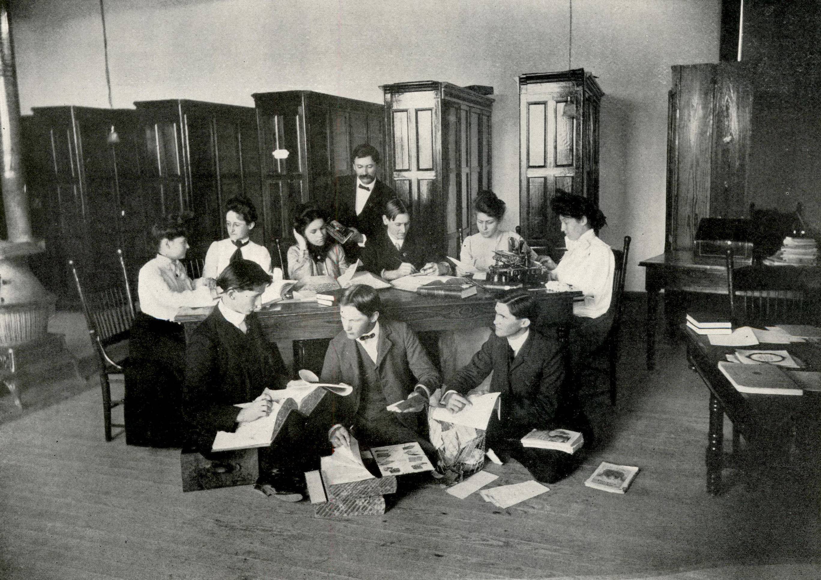 A group of students gather around a table in the library with gorgeous wooden bookshelves in the background. A professor looks onward as six students sitting at the table take notes, scroll through books, and share messages while three young men are huddled closely on the floor, researching a topic. All the students are dressed in Edwardian era clothing.