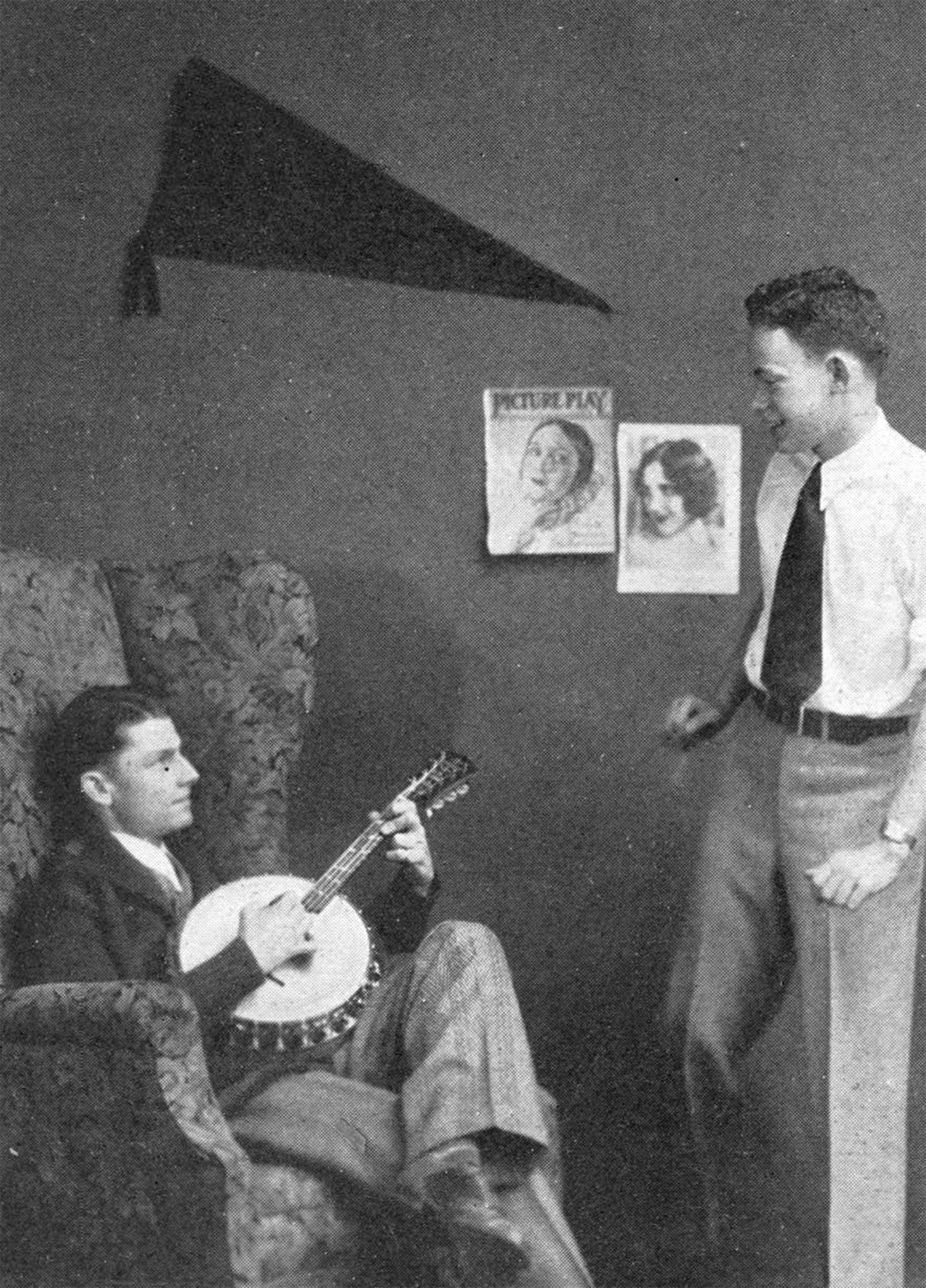 Candid aged black and white photograph of a pair of male students standing in their dorm room. One young man is seated in a high-backed upholstered chair with a floral pattern, smiling slightly while playing a banjo and looking up at his companion, standing nearby and looking down at his friend, mid-conversation. Decorating the wall behind them is a dark pennant and a few magazine clippings of pretty girls styled in the current flapper fashion with dark eye makeup and short hair.