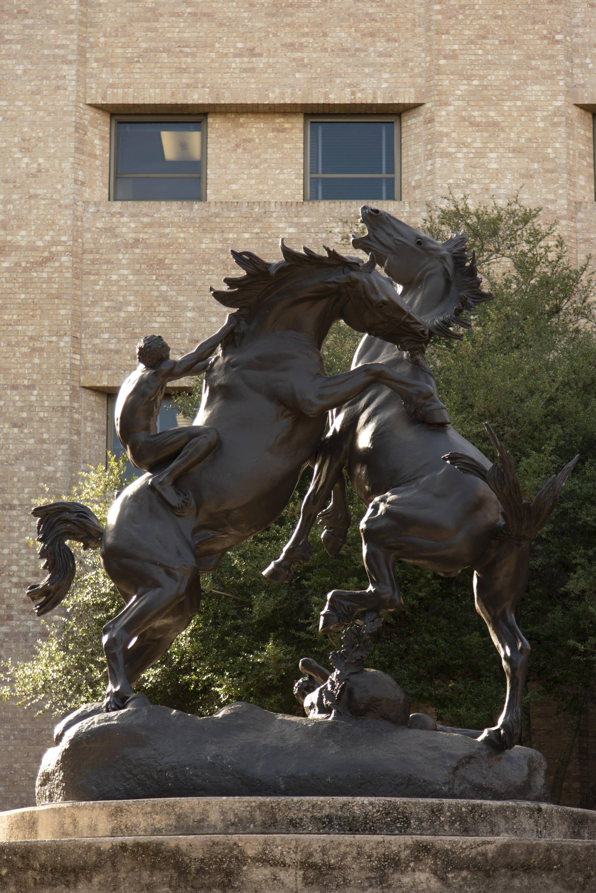 Photograph of the Fighting Stallions sculpture. Two massive horses are depicted rearing up as they engage in a fight. On the back of one, a man clings to the stallion, riding bareback and gripping the horse's mane to try to hold on. On the ground beneath the skirmish, another man can be seen, having been thrown from the second horse already.