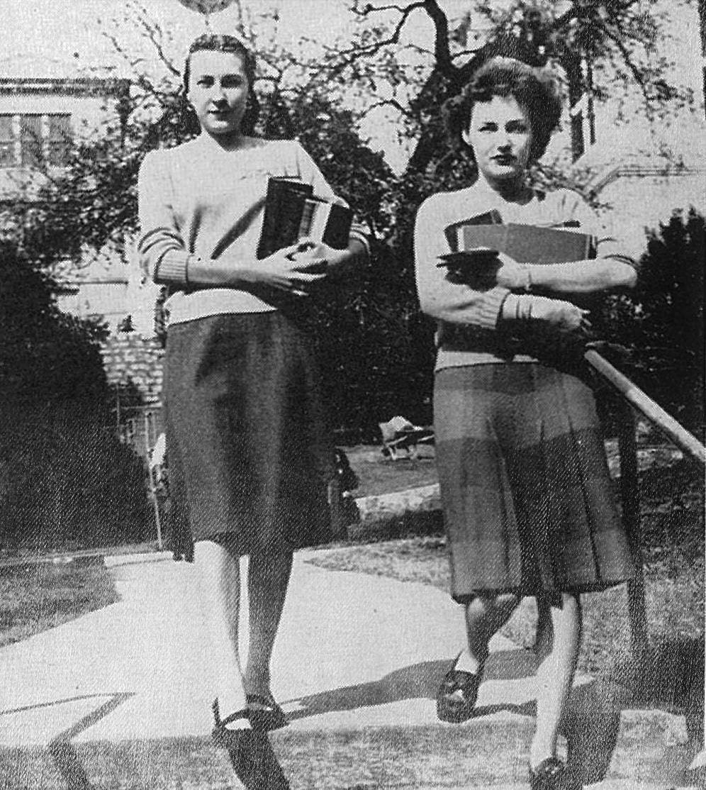 Vintage snapshot of a pair of female students walking across campus in 1947. Dressed neatly in sweaters and knee-length skirts, they're mid-stride, descending one of the campus' many steps as they juggle stacks of books in their arms.