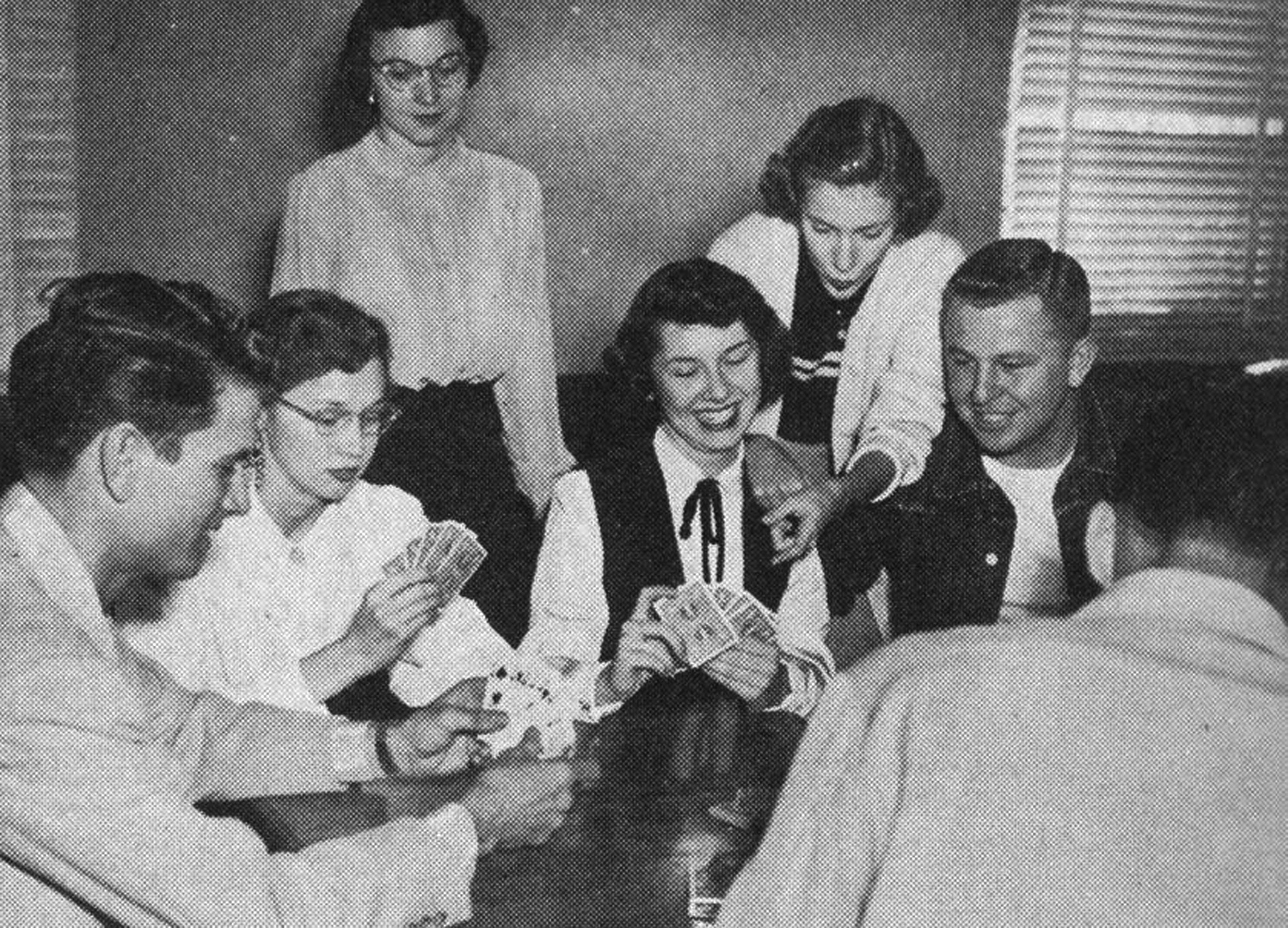 Black and white snapshot of a group of seven coed students playing cards in the 1950s. The camera looks over the back and shoulder of a young man. To his left, another young man and woman both examine their cards. Standing behind them, a lady watches with mild interest. Across the table, A young lady smiles as she plays one of her cards, and another woman standing behind her leans over her shoulder to gesture to her friend's hand. The last young man is to the smiling woman's left, and happily watches the game.