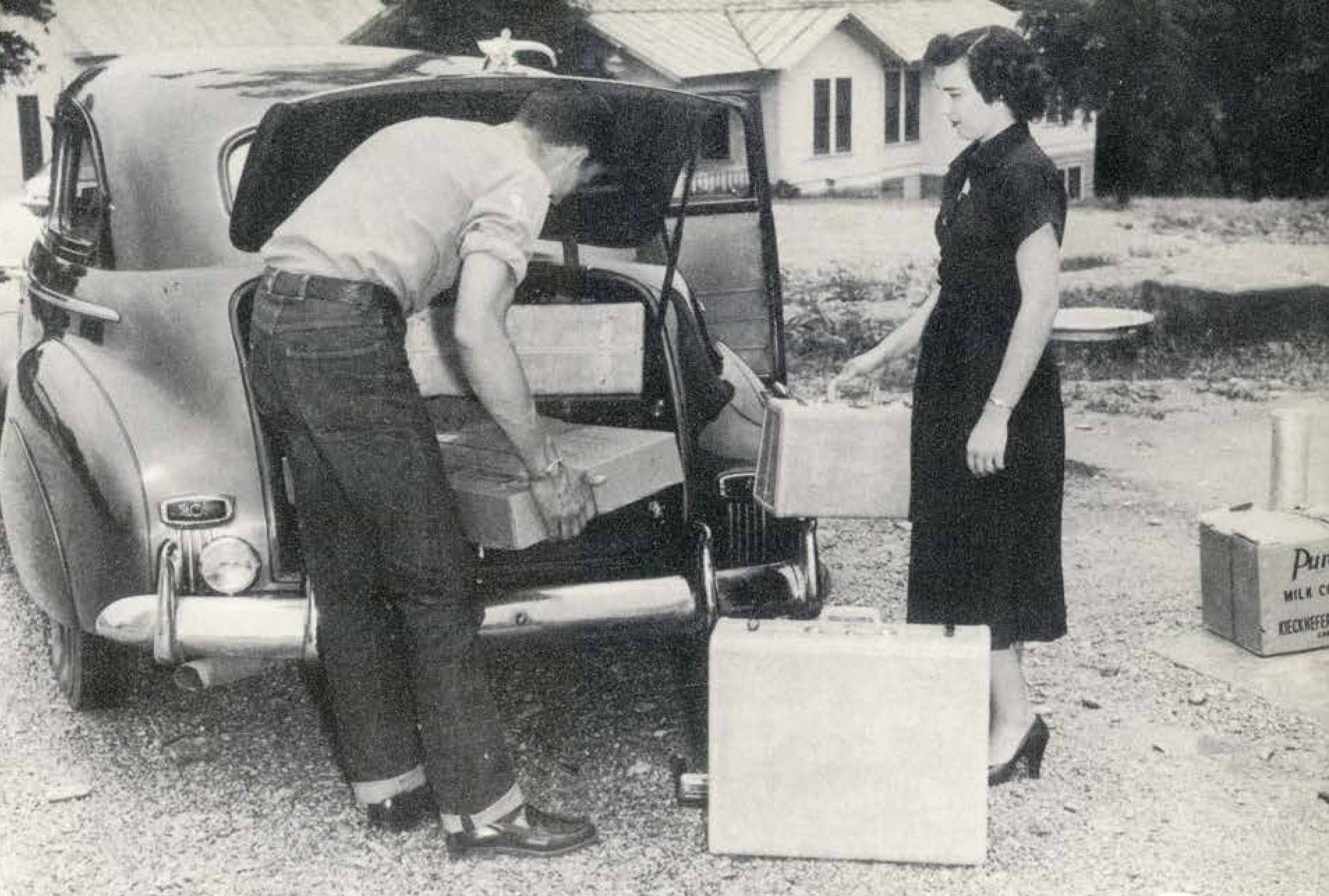 Black and white candid photo of man and woman unloading luggage to move one of them into the college dormitories. The young woman dressed in a dark tea-length dress with short sleeves and heels holds up a small suitcase, with a larger one set on the ground, watching while the man — wearing cuffed jeans and a shirt with his sleeves rolled up — stoops to lift a large box out of the trunk of an old station wagon.