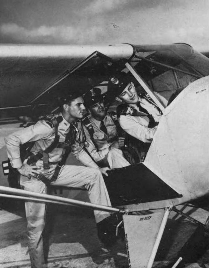 Vintage snapshot of a trio of Air Force ROTC cadets around a small plane. Two are seated in the plane's cockpit, the man in front showing the other two something on the plane's controls. The second man, seated behind the first, leans up to see what the first is talking about, and a third man leans on the step on the plane's step-up, listening.