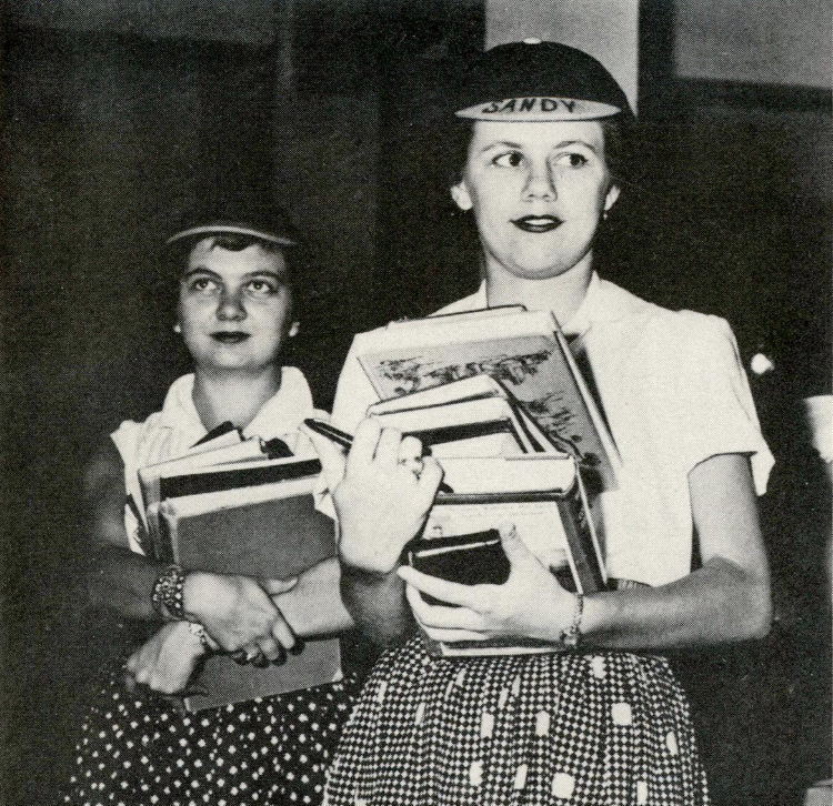 Candid photo of a pair of freshmen girls wearing the traditional freshman beanie. They're dressed in skirts and light shirts, and each holds a stack of books. The beanies are dark caps with a small, short brim in a contrasting color. One girl's beanie has her name written across the top of the brim, 'Sandy.'