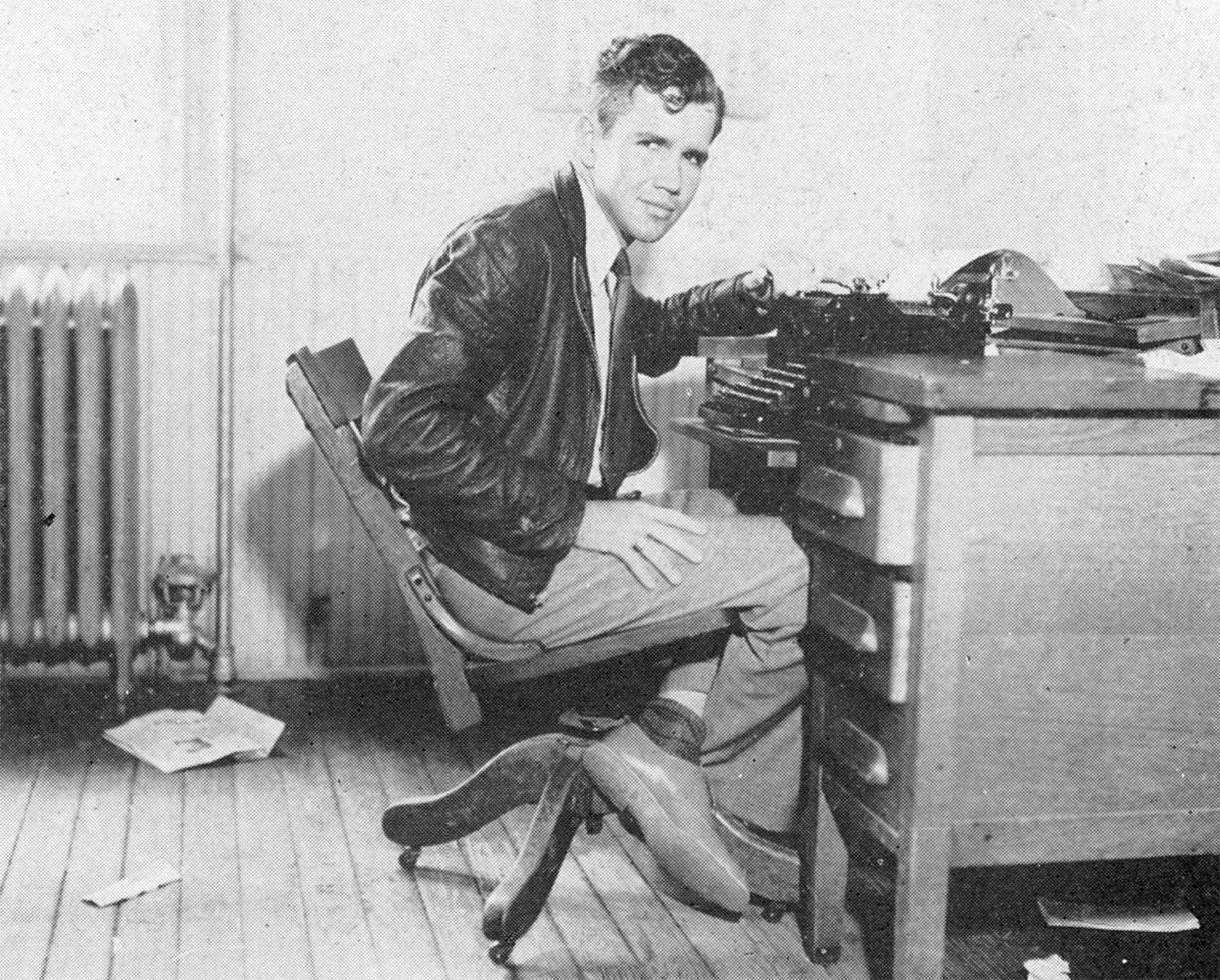 Vintage black and white photograph of a young man seated at a heavy wooden desk, with a bit of paper clutter scattered around. Shown in profile, he's dressed in a dark leather jacket, collared shirt with a neck tie and slacks. He's turned his head to face the camera with a slight smile as he leans his wrist against a typewriter on the desk.