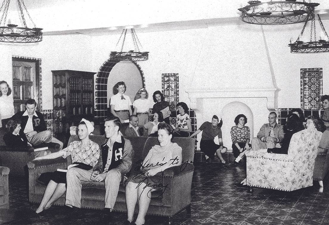 Aged black and white snapshot from the 1948 Pedagog, of a student community space. The room is decorated with fixtures done in the Spanish revival style, with a patterned tile floor, and dark tiles going halfway up the wall. A pair of wall niches decorated with patterned tile frame a large fireplace mantel. To the right of the fireplace, a tiled arch leads to another room, and a heavy wooden bookcase stands in the corner next to a window decorated in the same tile as the wall niches. Several pieces of comfortable furniture are placed in the room, including a dark sofa with matching armchairs and a floral armchair facing the fireplace. Simple wrought iron chandeliers with candle flame-shaped bulbs light the room. In the room, co-ed students are mulling around and socializing. On top of the photograph, someone has scrawled 'Here's to you, Nancy.'