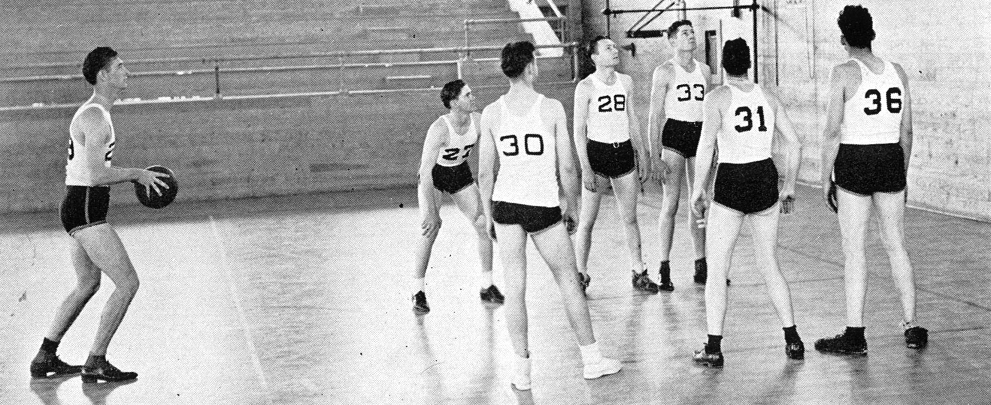 Vintage black and white photo of a group of young men playing basketball in a gymnasium. Wearing dark shorts and numbered jerseys, six of the men are lined up watching the basket as a seventh man dribbles and prepares to make a freethrow shot.