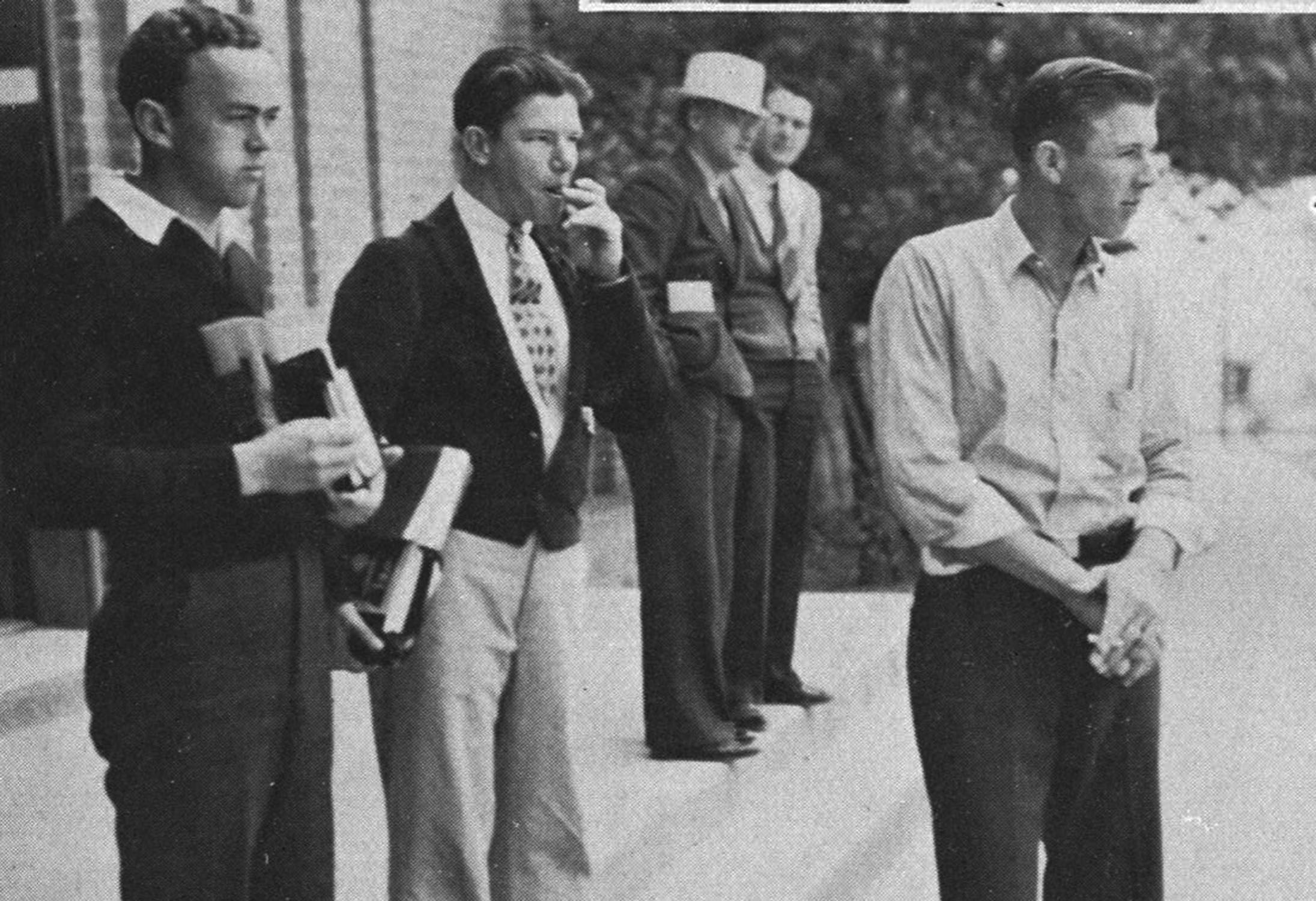 Candid aged black and white photo from 1937. A group of men are all standing on the stoop of a brick building. Three young men stand in the foreground, holding their books while having a smoke break before carrying on with their day. One wears a university sweater with a large 'T,' one wears a neat suit with a patterned tie and short jacket, and the third wears slacks and a button down shirt, with collar open and the sleeves rolled up casually. Behind them, two more older men, one in a suit and hat, the other in a cardigan and neck tie, stand chatting with their hands in their pockets. All the men are facing the right, and appear to be casually observing some sort of occurrence outside the image frame.