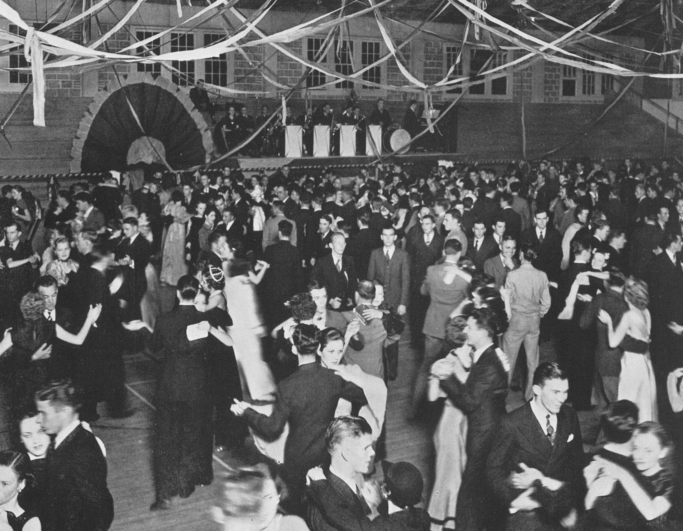 A vintage black and white photograph of a school dance in 1937. A band plays on a dais in the background set up against the far wall. Streamers have been stretched above a large dance floor, crowded with young men and women as they dance the night away.