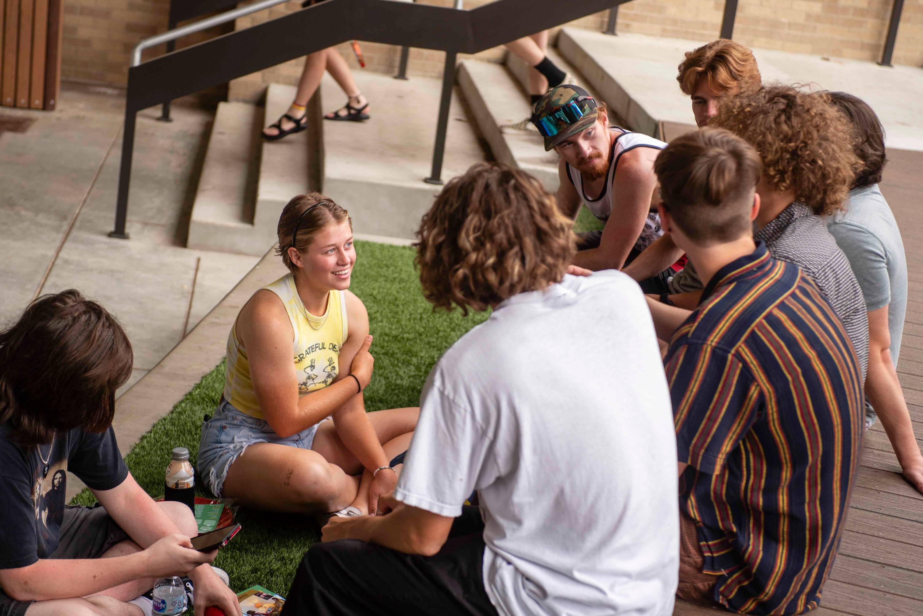 A gaggle of young people sit in the grass, chatting.