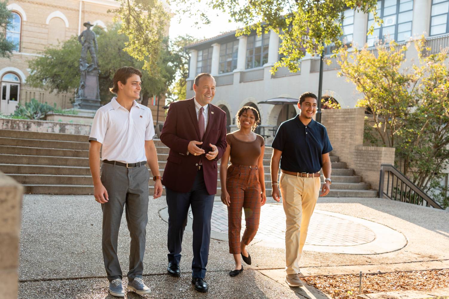 A man in a suit talking and laughing with three young people down a sunlit brick path.