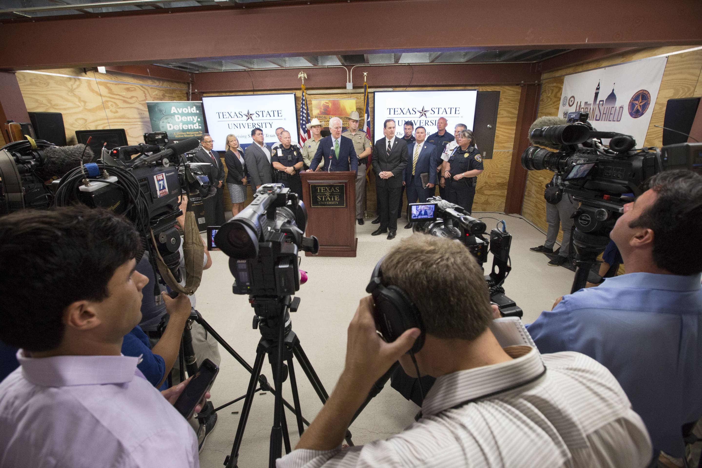 A shot over the shoulder of camera men who are in turn shooting a press release. A man in a suit speaks at a podium and behind him are other people in suits and several uniformed police officers.