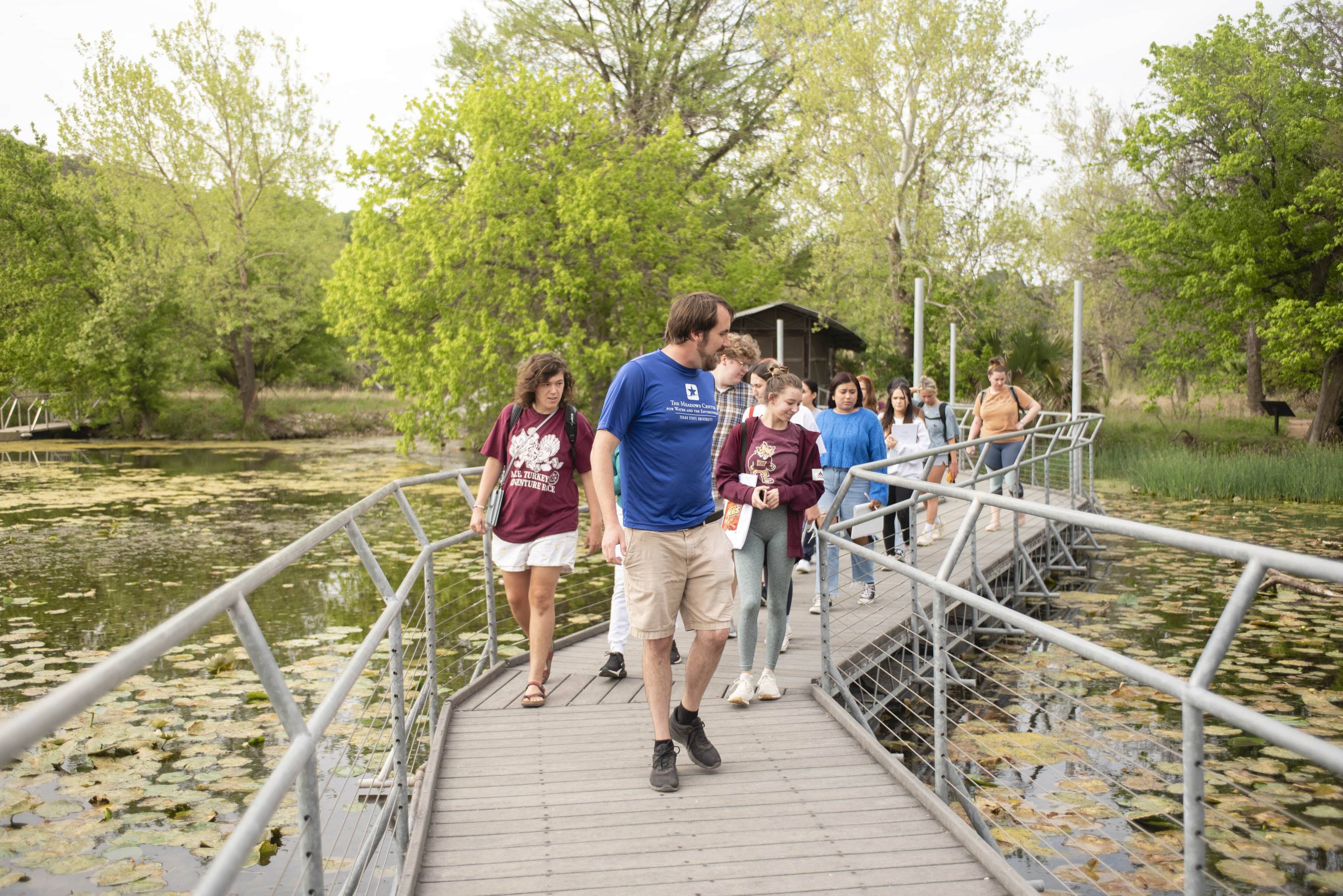 A man in a blue T-shirt leads a group of onlookers over a boardwalk. On either side is water covered in lilypads, which captures the attention of the onlookers. In the background trees can be seen.