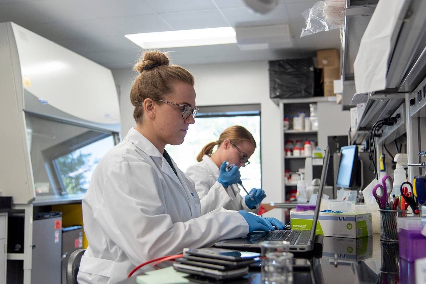 Two young women in a laboratory wear white labcoats and blue gloves. The woman in the foreground types on a laptop while the woman in the background is extracting liquid from a vial. 