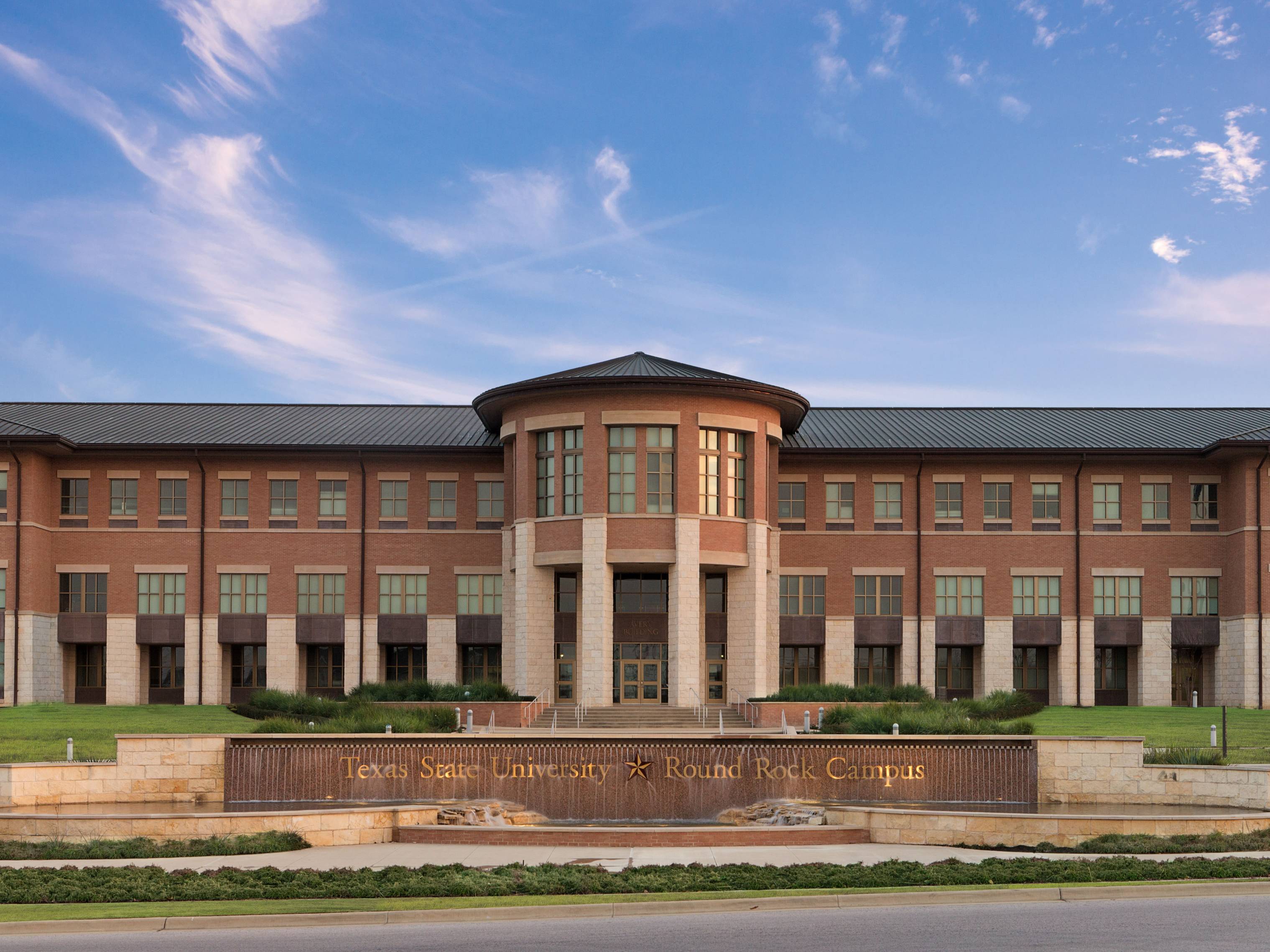 A stone and metal signs says "Texas State University -- Round Rock Campus". Behind it, an imposing stone building is awash in the light of dusk.